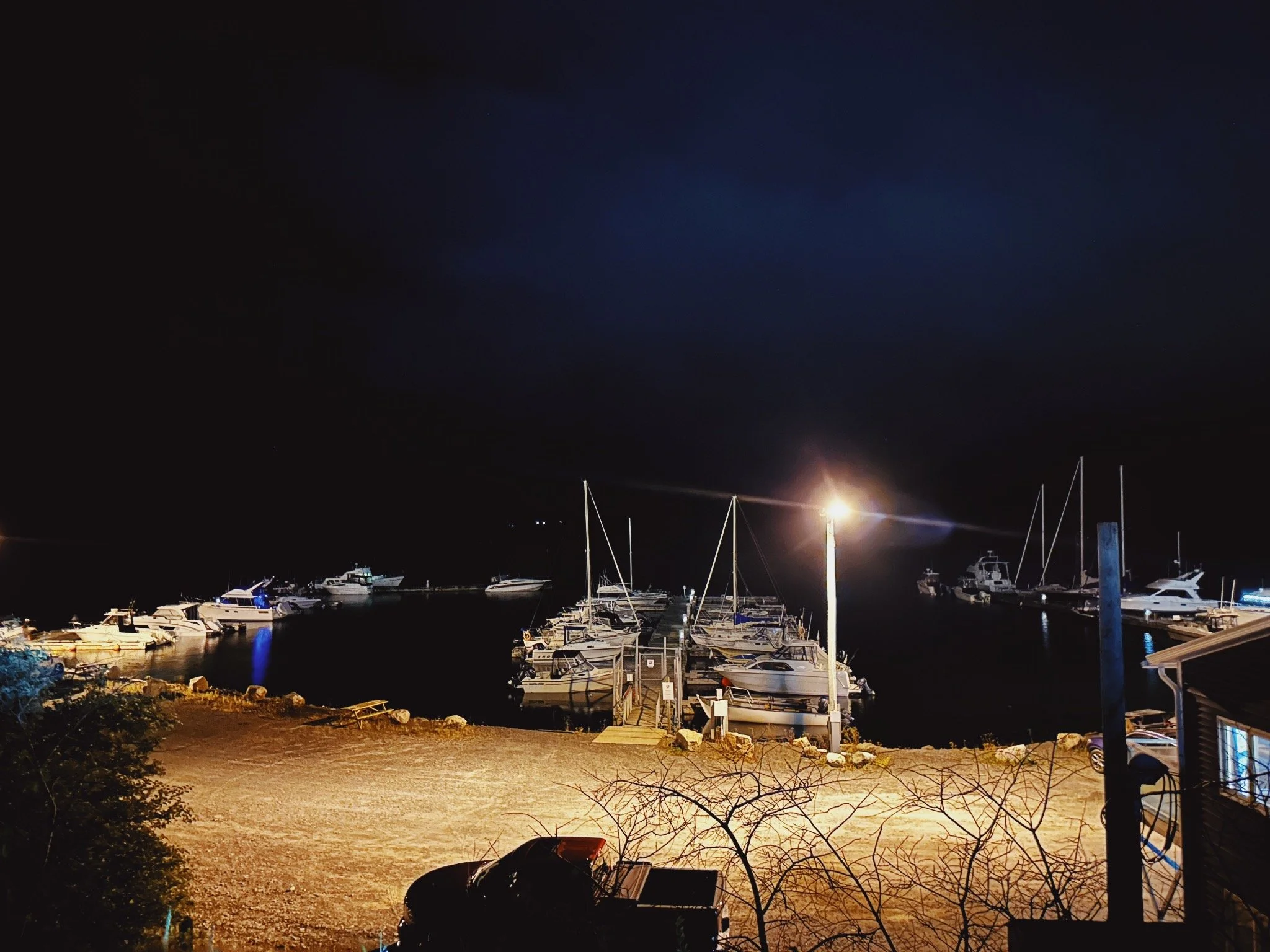 Nighttime view of boats docked at a marina with masts visible, illuminated by a bright light, under a dark sky.