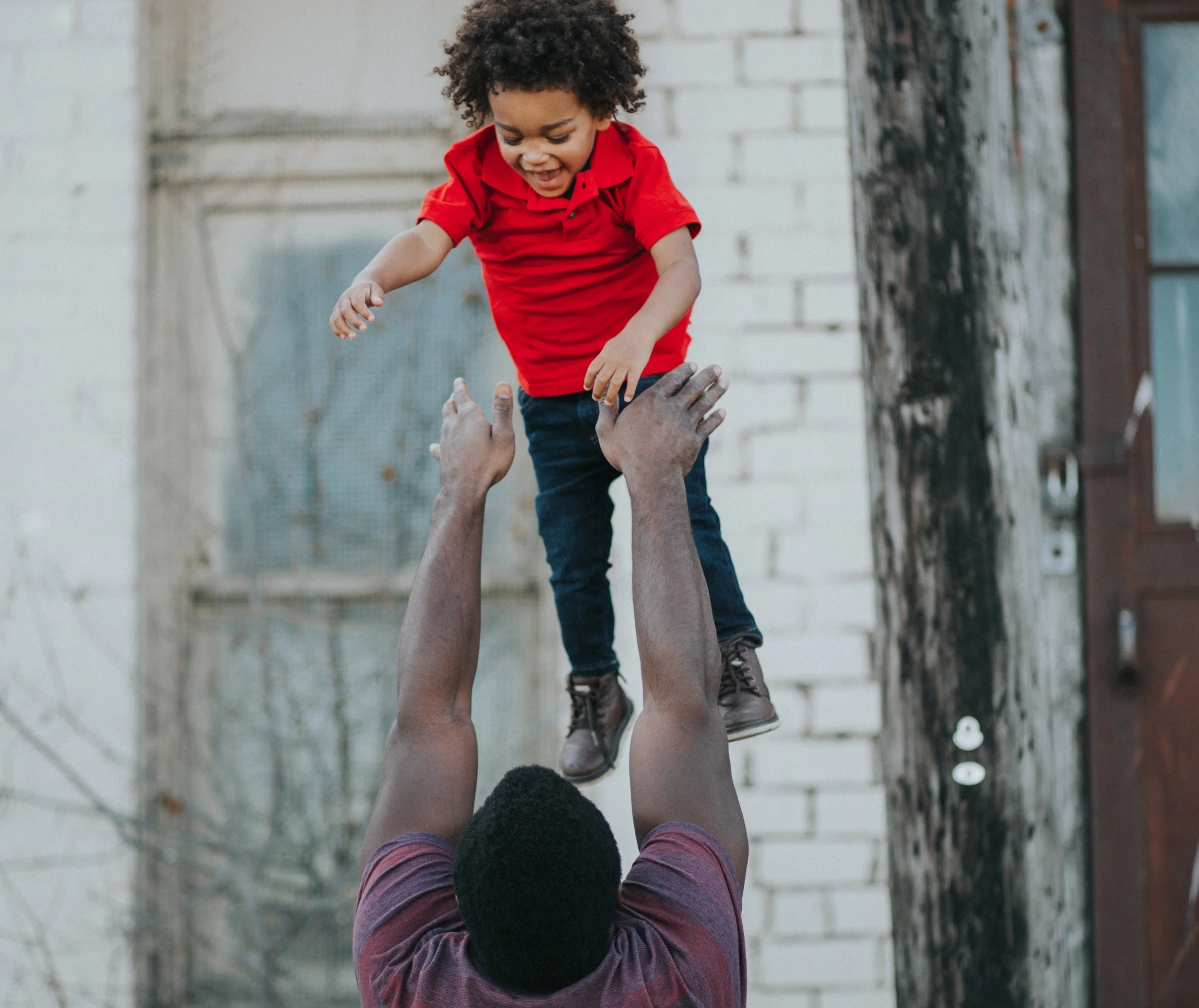 A young boy with curly hair and a red shirt being lifted in the air by an adult with dark hair and a striped shirt, against a backdrop of a white brick wall.