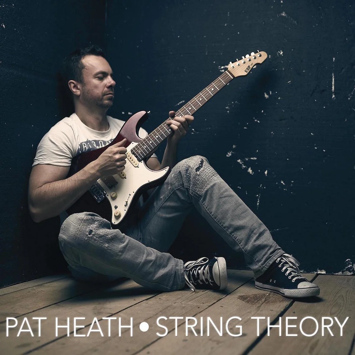 A man sitting on a wooden floor against a black wall, playing an electric guitar.