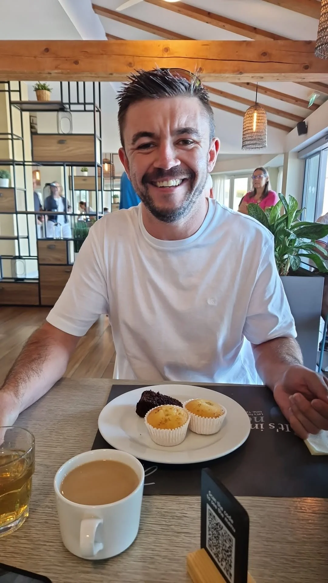 A smiling man in a white t-shirt sitting at a table in a well-lit restaurant, with a plate of desserts including a piece of chocolate cake and two small tarts, along with a glass of what appears to be tea or coffee, and a cup of tea or coffee in front of him.