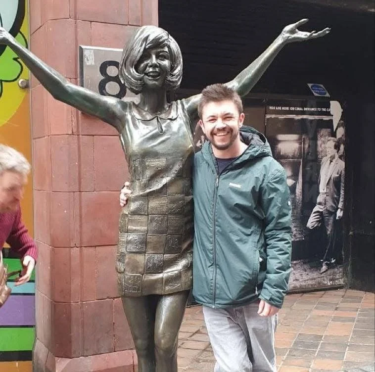 A man smiling and posing with a life-sized statue of a woman with arms raised, outside a building with brick walls.
