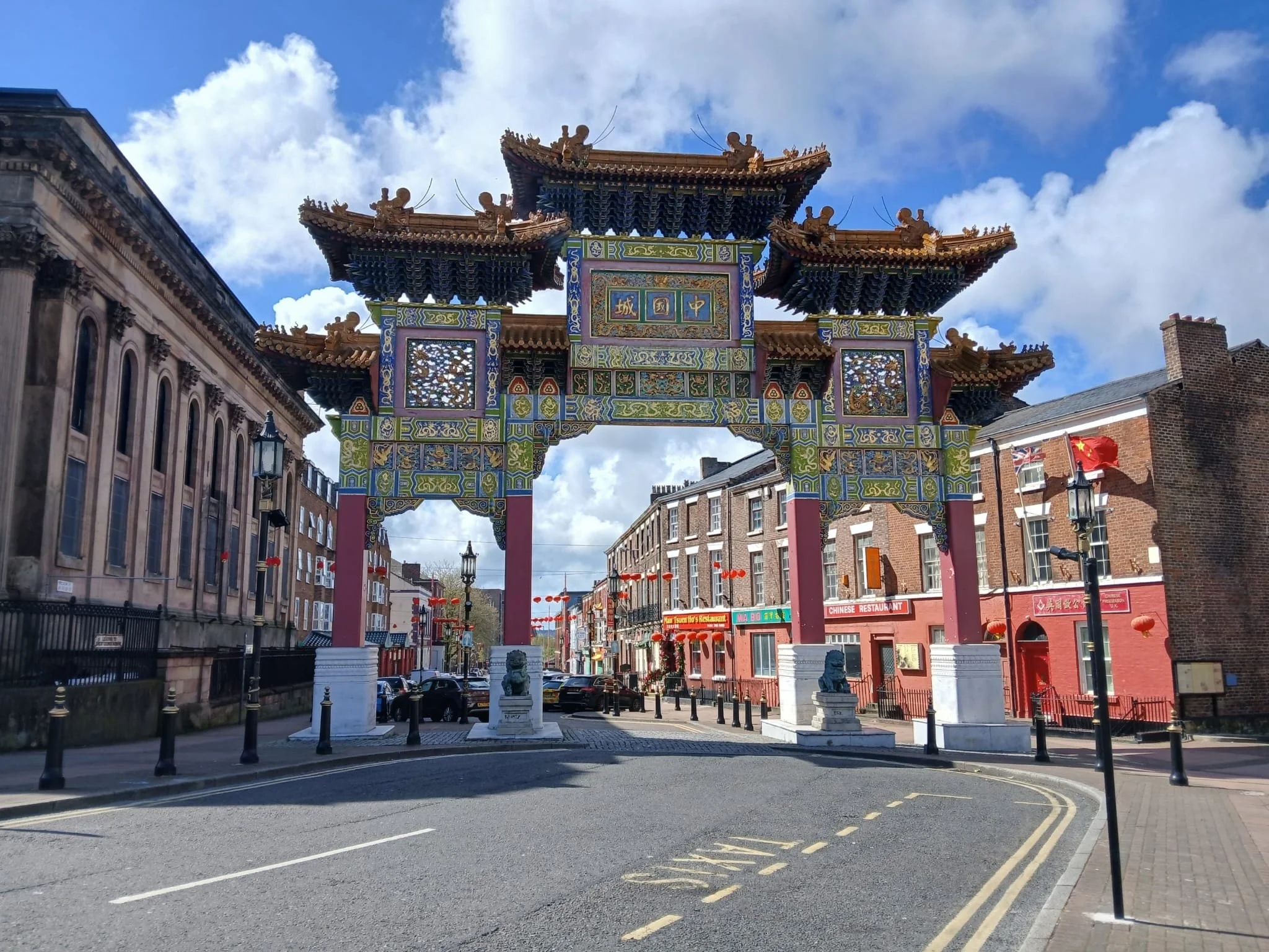 Street scene with a large, colorful, traditional Chinese gate in the center, surrounded by buildings with brick and stone facades, red lanterns hanging, and blue sky with clouds overhead.
