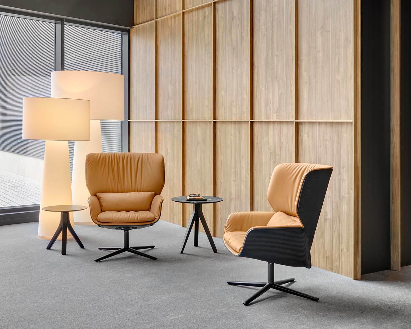Modern office interior with two Andreu World chairs, a small round Andreu World black side table, a floor lamp, and a wooden wall paneling, next to large windows with horizontal blinds.