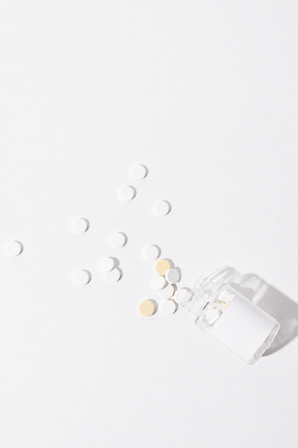 White and yellow pills spilled from a small overturned glass bottle onto a white surface.