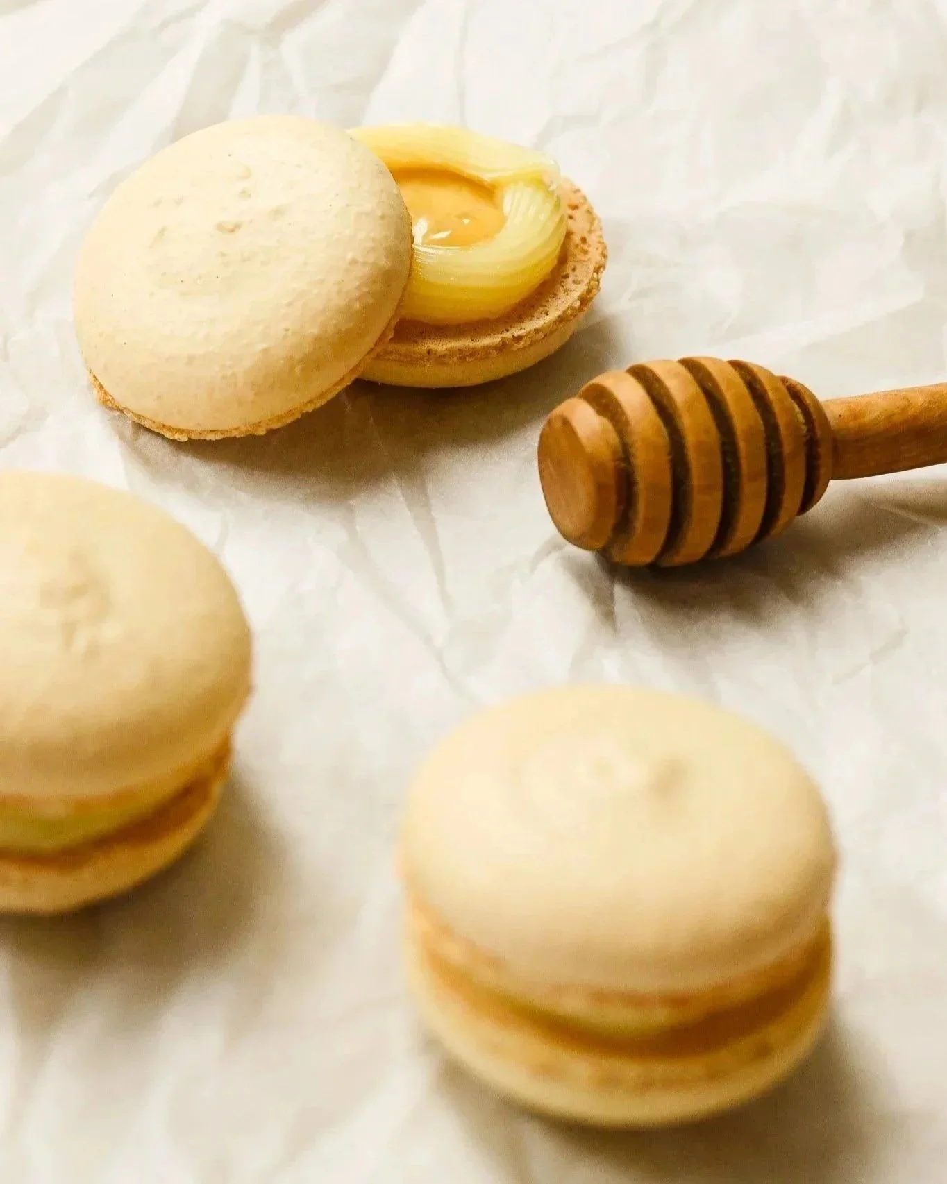 Close-up of macarons with a honey dipper on white parchment paper, with one macaron cut open showing creamy filling and onion slices inside.