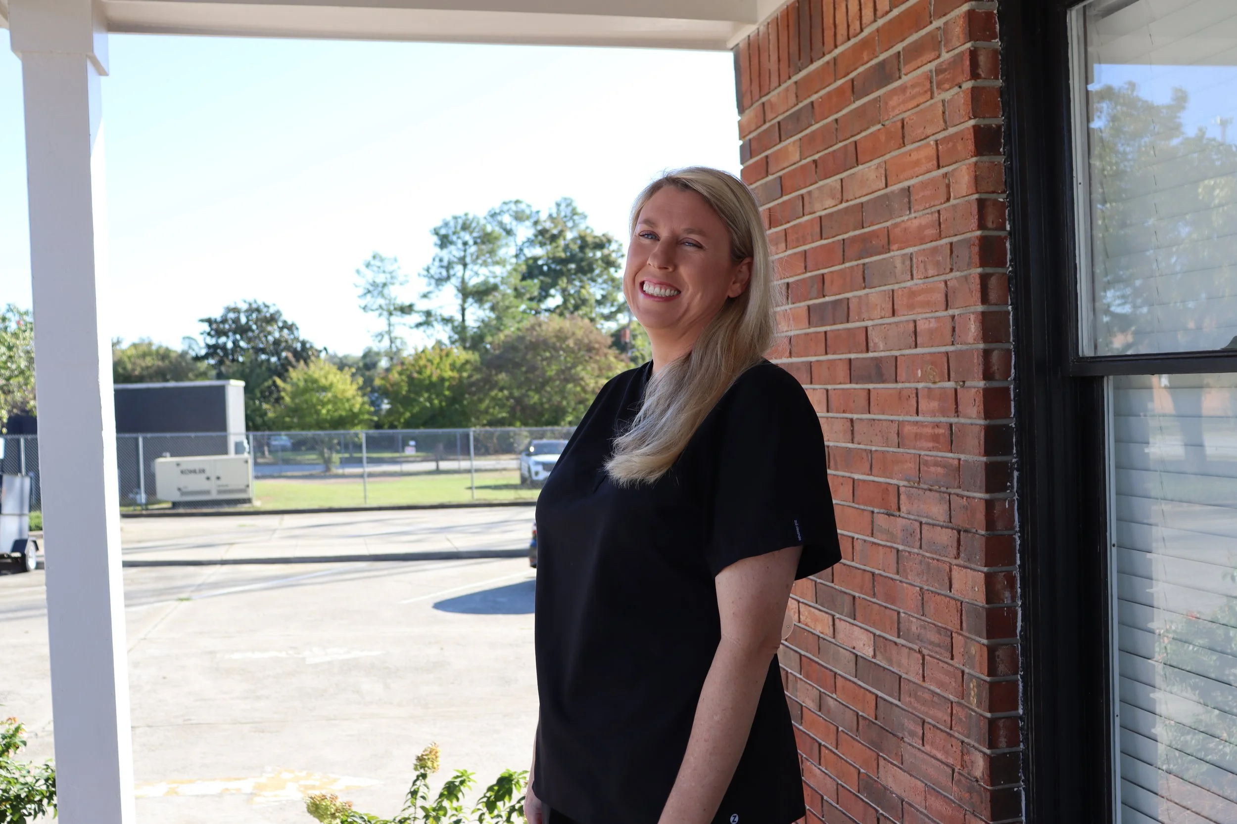 A smiling blonde woman standing outside near a brick wall, with trees and a parking lot in the background.