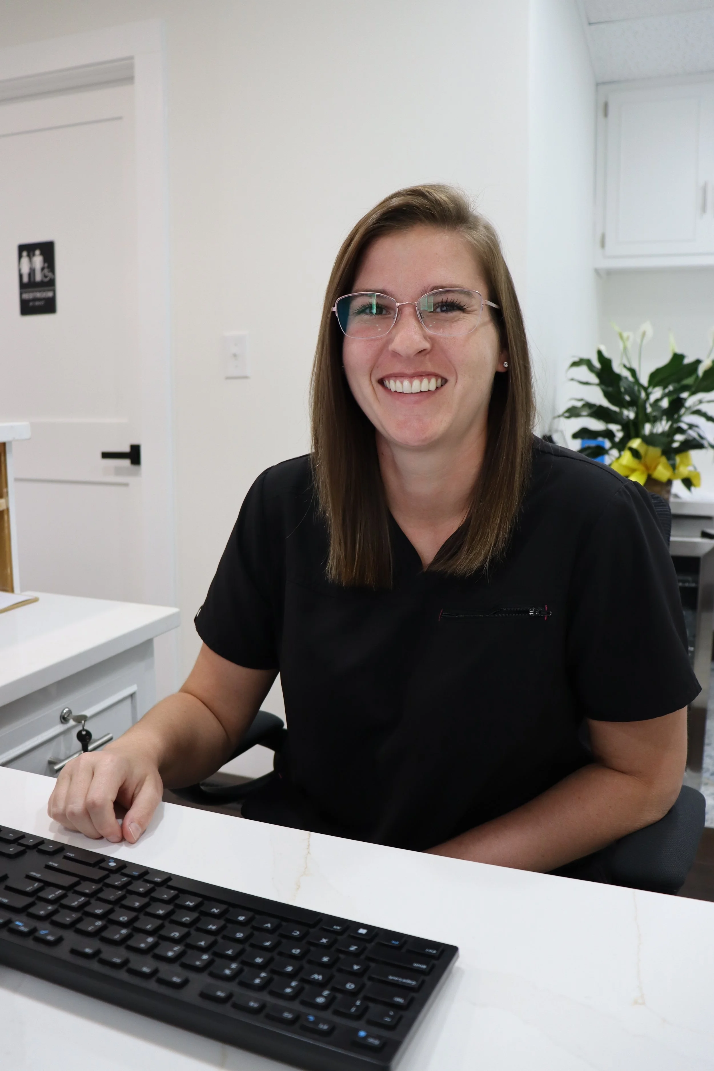 A woman with shoulder-length brown hair, glasses, and a black top sitting at a desk smiling at the camera. A computer keyboard and white desk are in the foreground, and a potted plant with white flowers and a yellow bow is in the background.