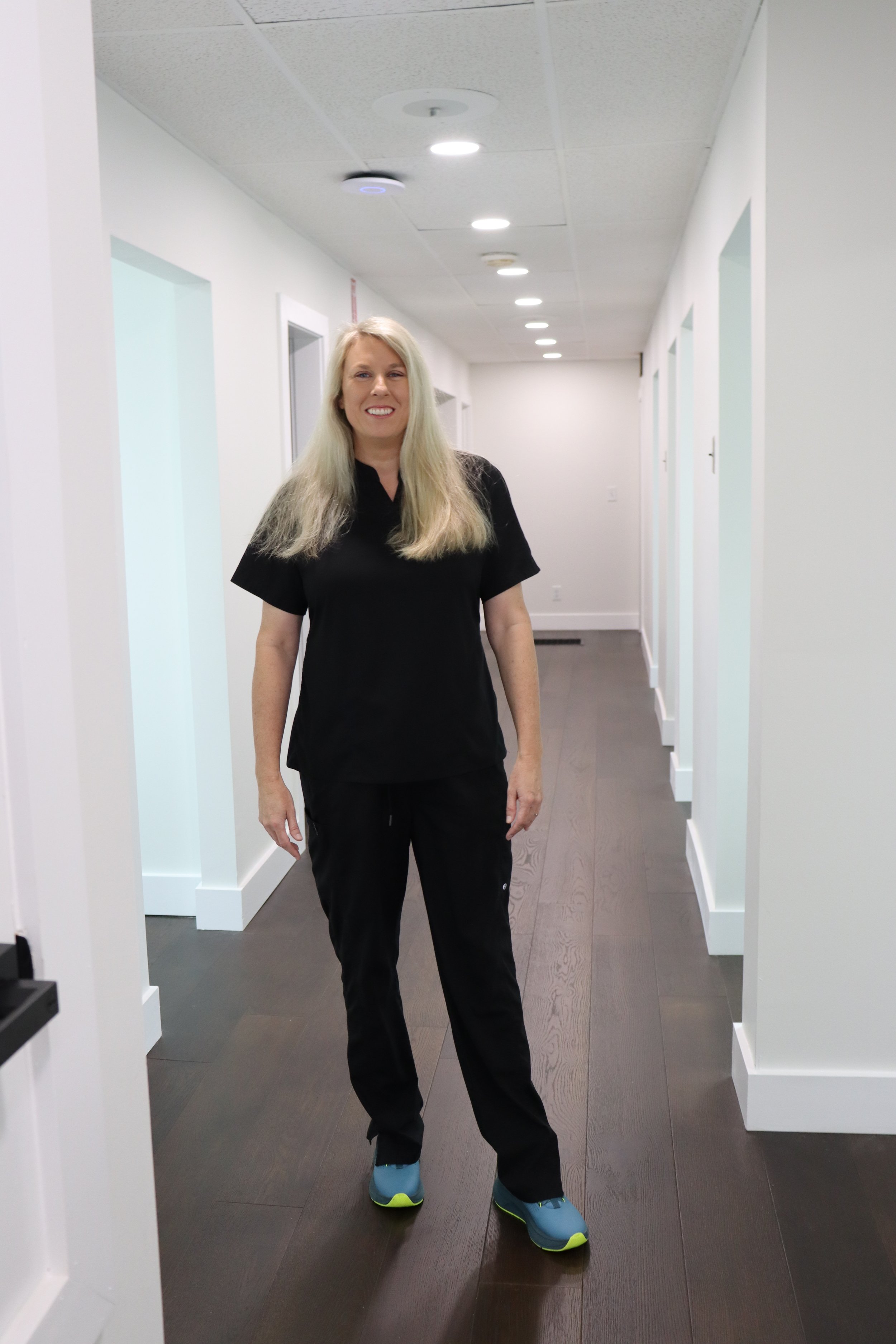 A woman with long blonde hair wearing a black scrub top, black pants, and blue sneakers, standing in a brightly lit corridor with white walls and dark wooden flooring.