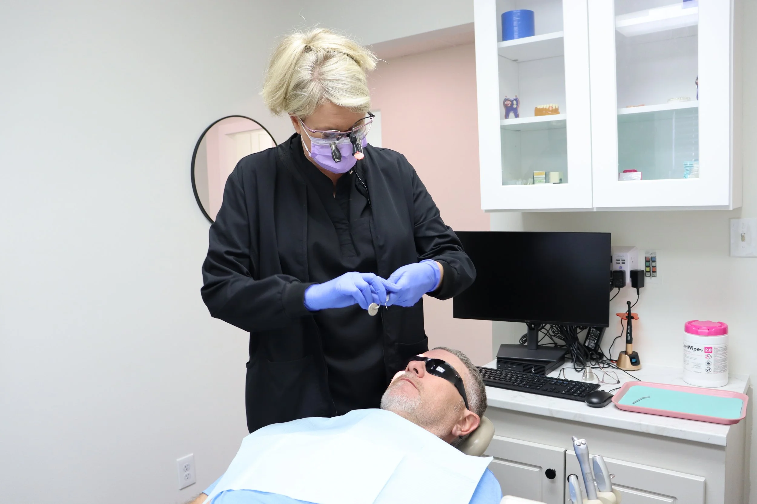 Dental professional wearing magnifying glasses and a face mask attending to a patient lying on the dental chair in a clinic. The clinic has white cabinets with dental supplies, a computer monitor, and various dental equipment on the counter.