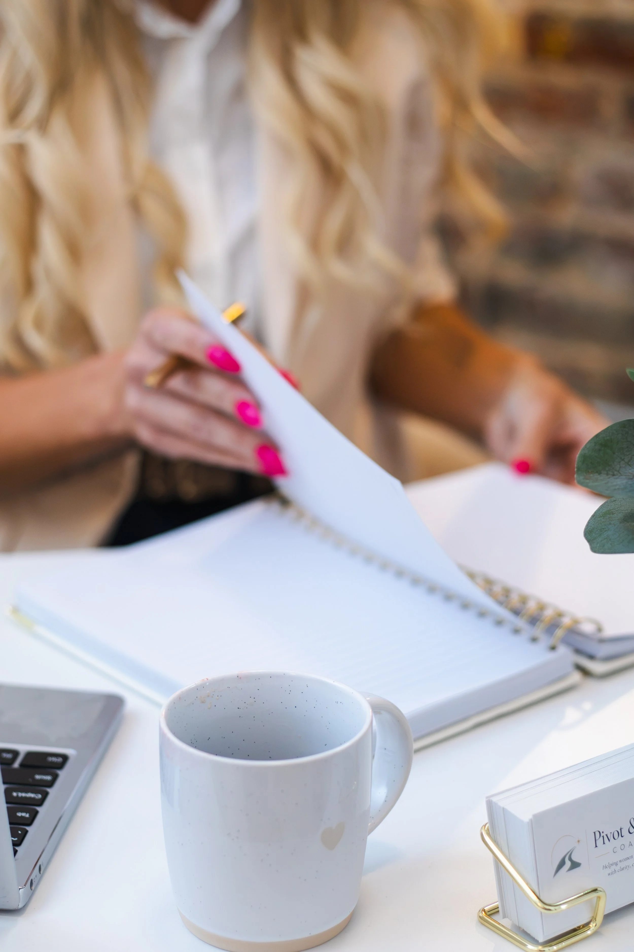 A white ceramic mug with a small gold heart on it, filled with a dark beverage, is on a white desk. Behind the mug, there's an open notebook, a laptop, and a cup of coffee or tea. A woman with long blonde hair, wearing a beige blazer, is holding a pen and writing in the notebook. There's also a small stack of business cards in a gold cardholder on the desk.