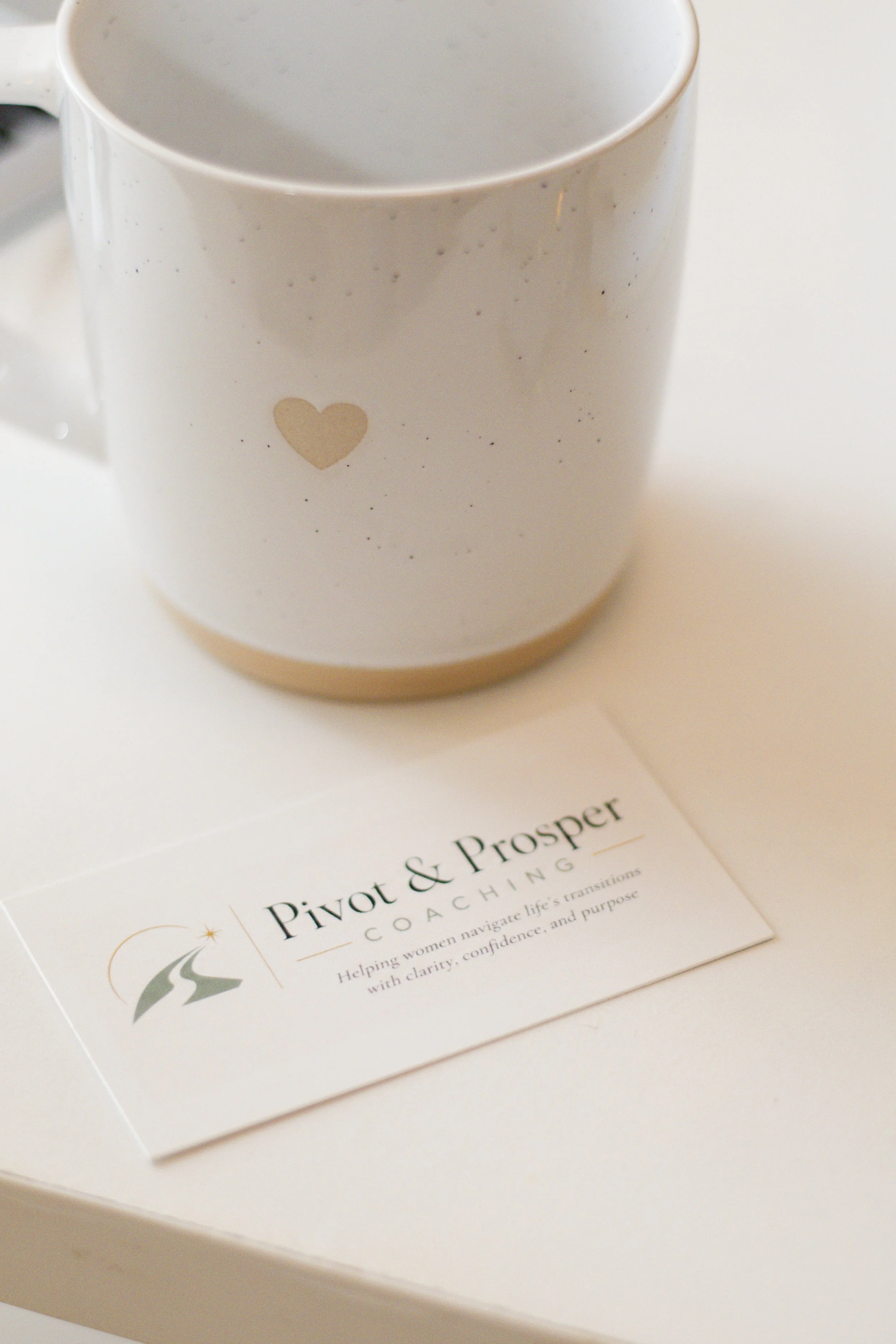 White mug with sparkly black speckles and a small beige heart on the side, placed on a white paper with black text that reads 'Pivot & Prosper.' The paper appears to be a business card or flyer for a coaching service.