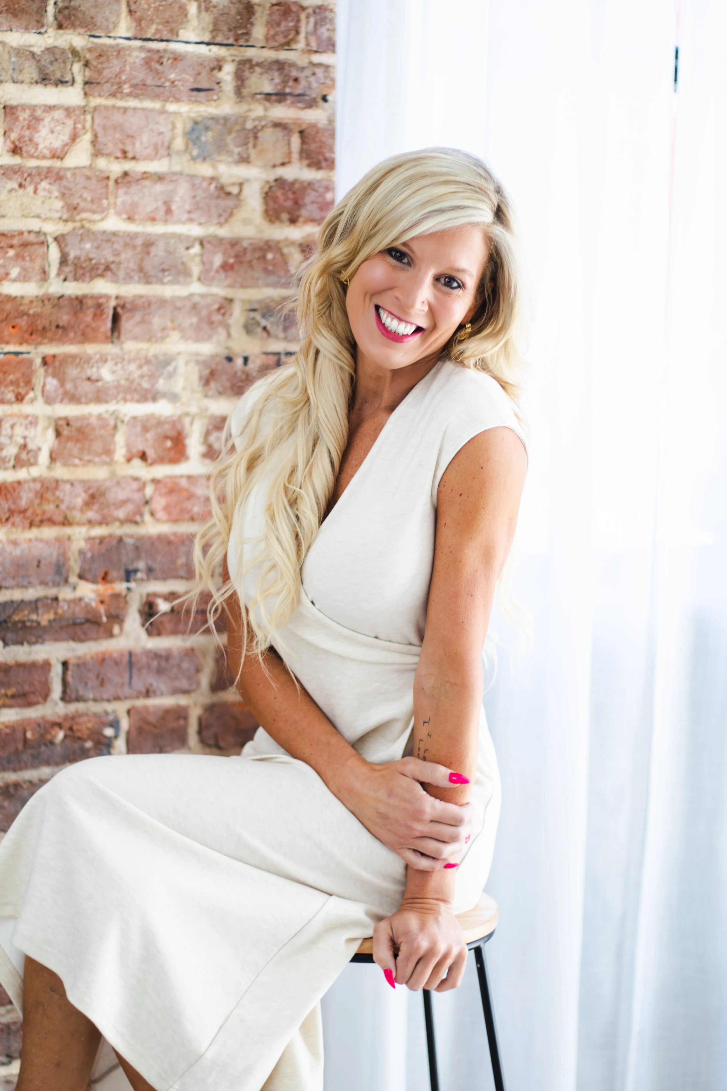 A woman with long blonde hair sitting on a black stool near a brick wall, smiling, wearing a white dress with rolled-up sleeves, and bright pink nails.