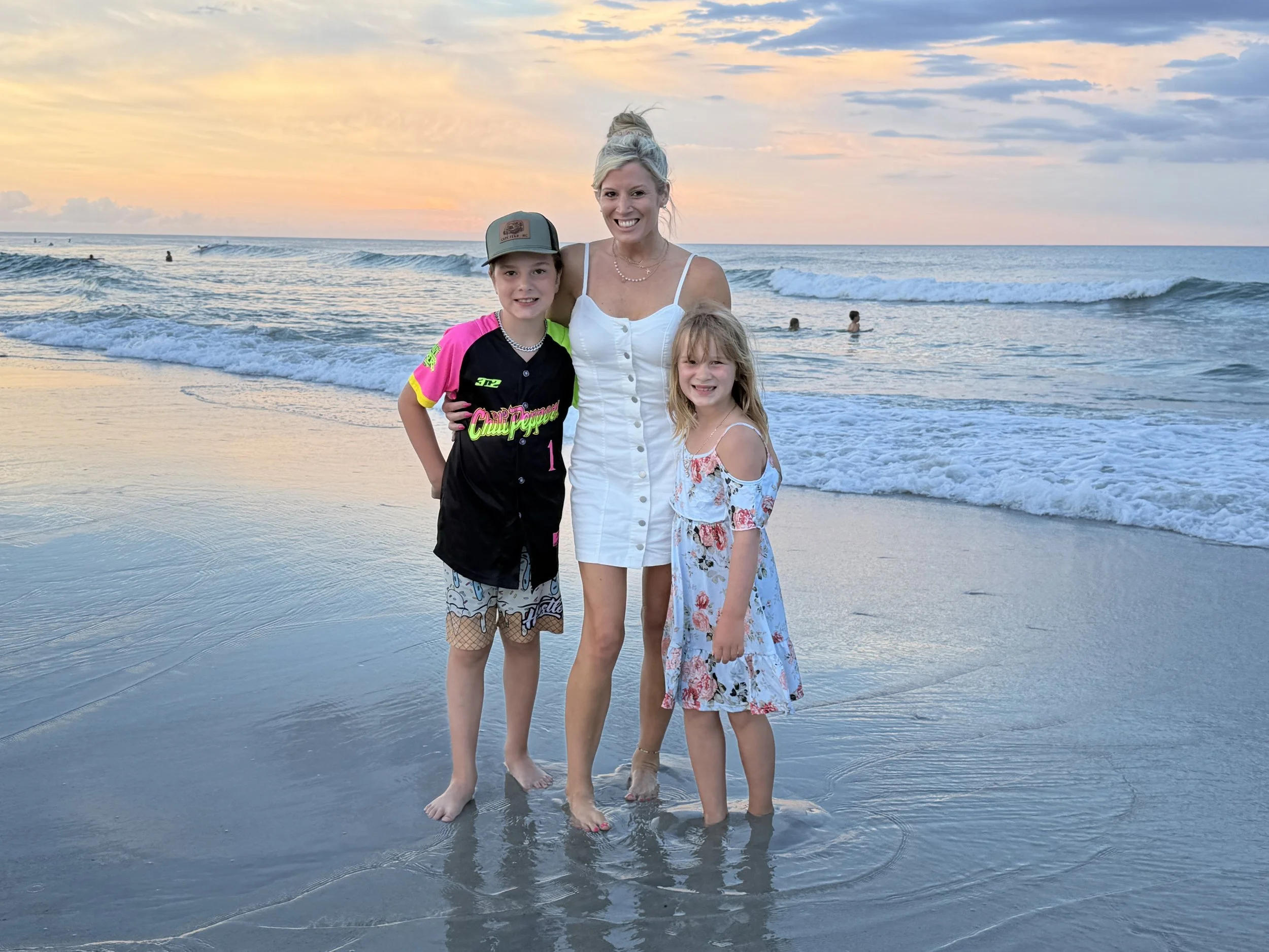 A woman and two children standing in shallow ocean water at sunset, smiling at the camera, with waves and surfers in the background.