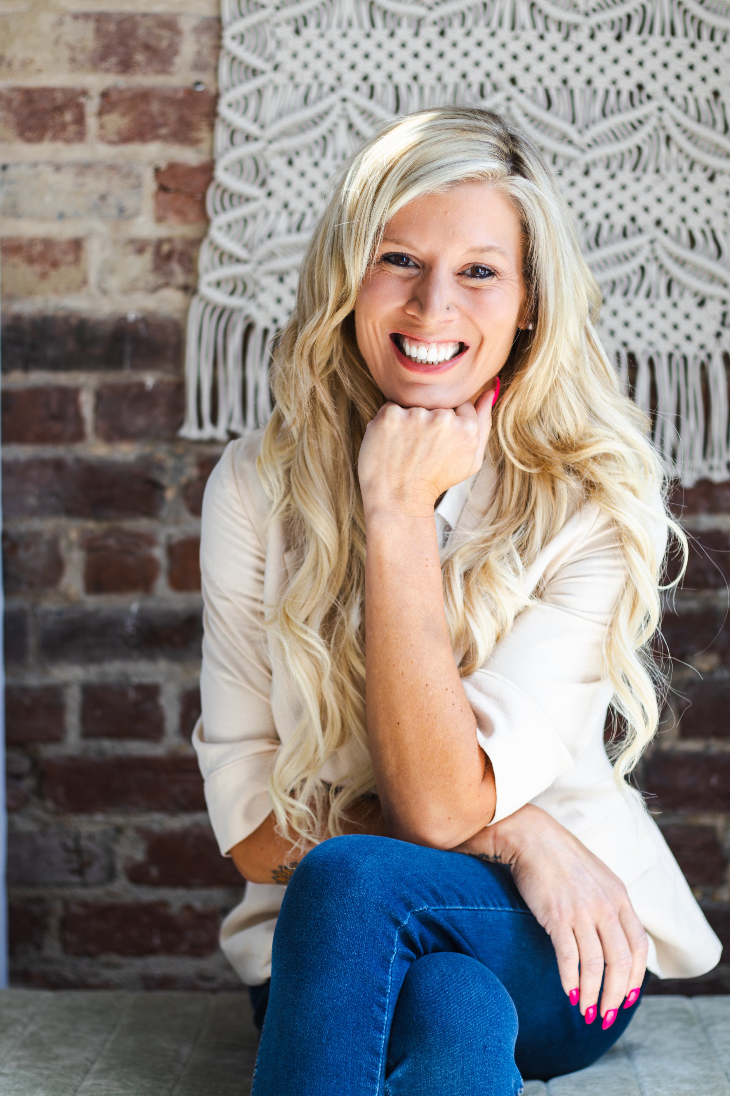 A smiling woman with long blonde hair, sitting with one leg crossed over the other, wearing a cream-colored shirt and blue jeans, against a brick wall with a white woven wall hanging in the background.