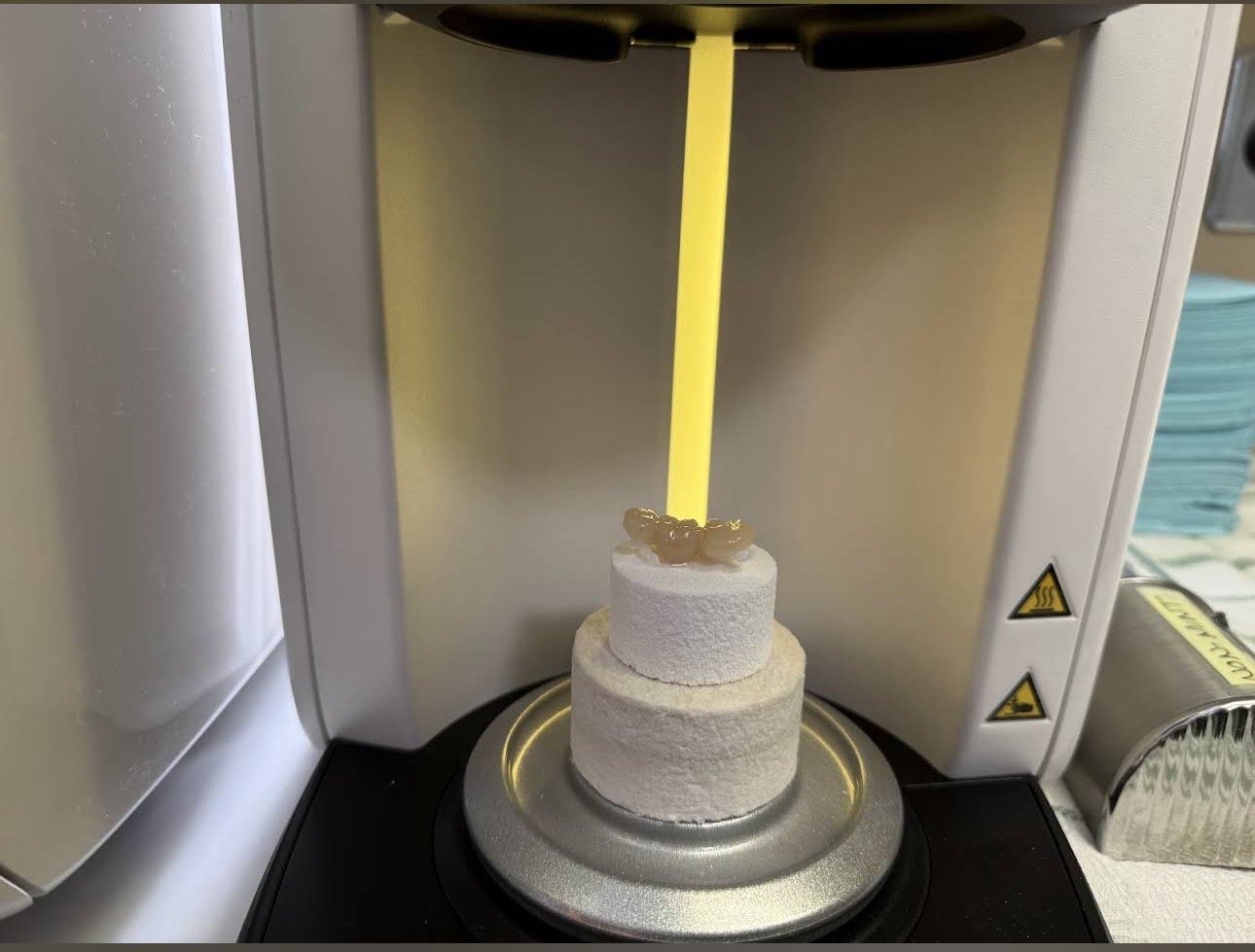 3D printer layer by layer creating a white cylindrical object with a small topping, inside a 3D printing chamber.
