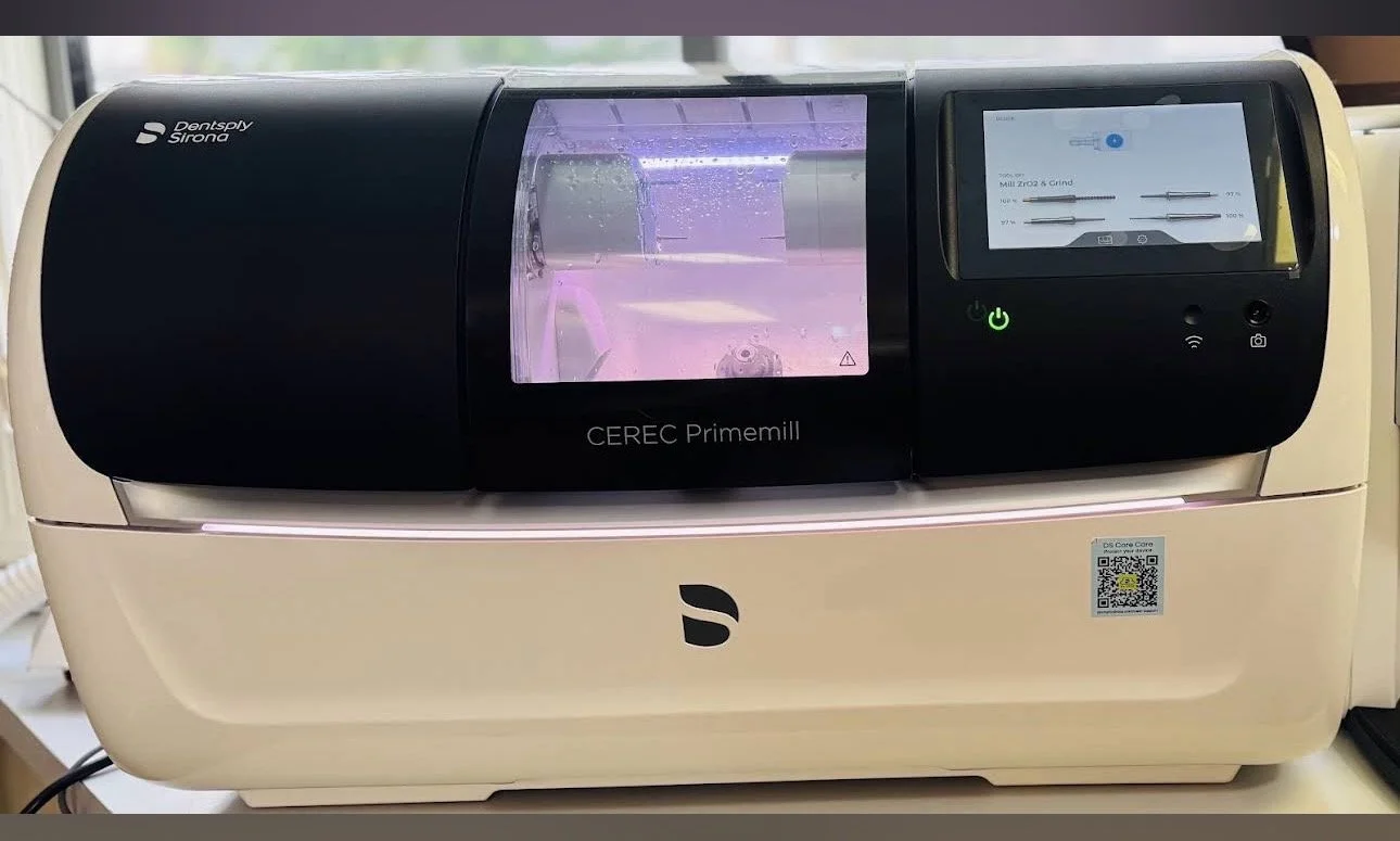 A medical laboratory device named CEREC Primemill from Dentsply Sirona with a digital screen showing settings, a transparent cover over the milling area, and a beige base with a QR code and logo.