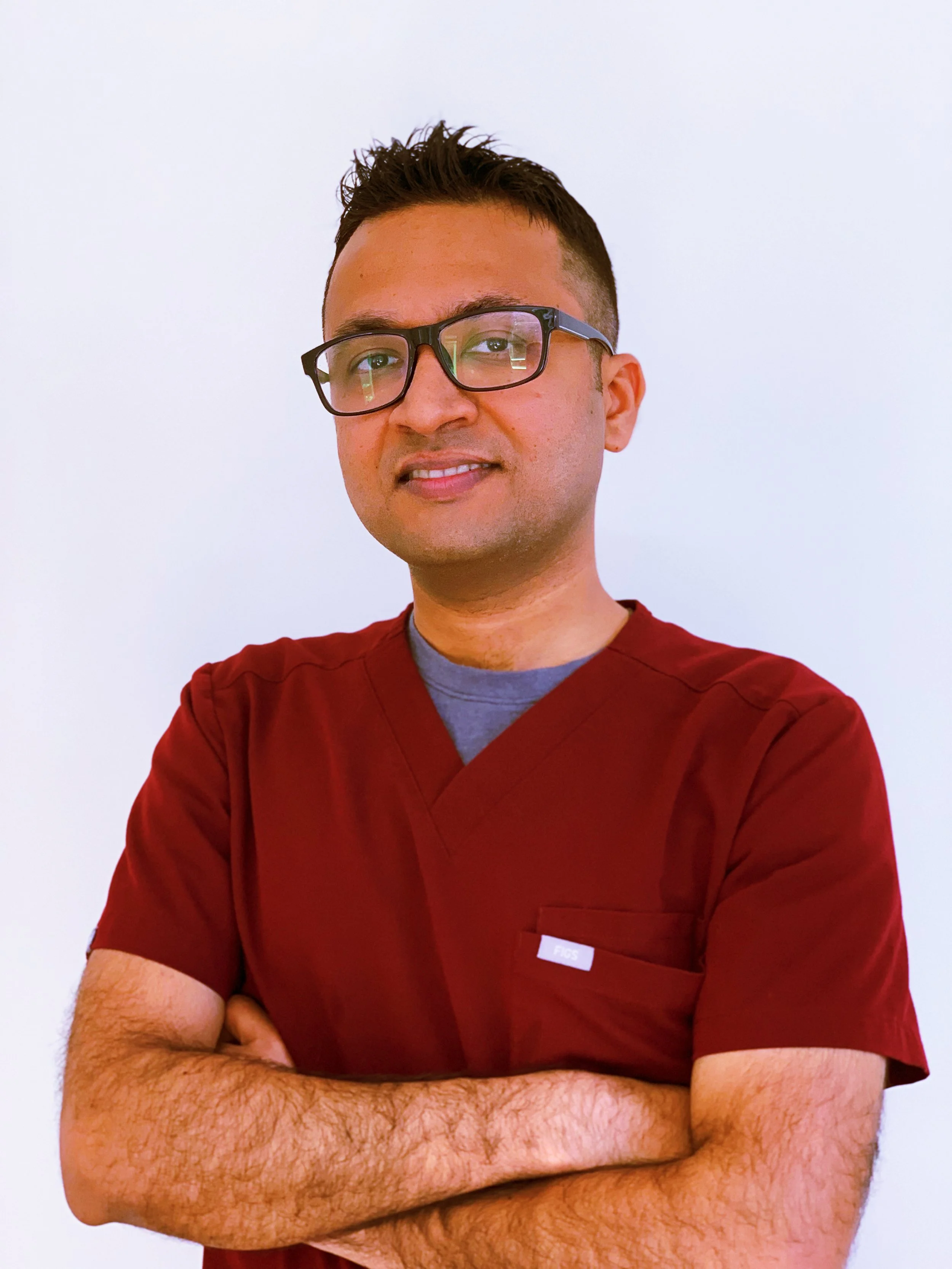 A man with glasses, short dark hair, and light skin, wearing a maroon medical scrub top, standing with arms crossed against a plain white background.