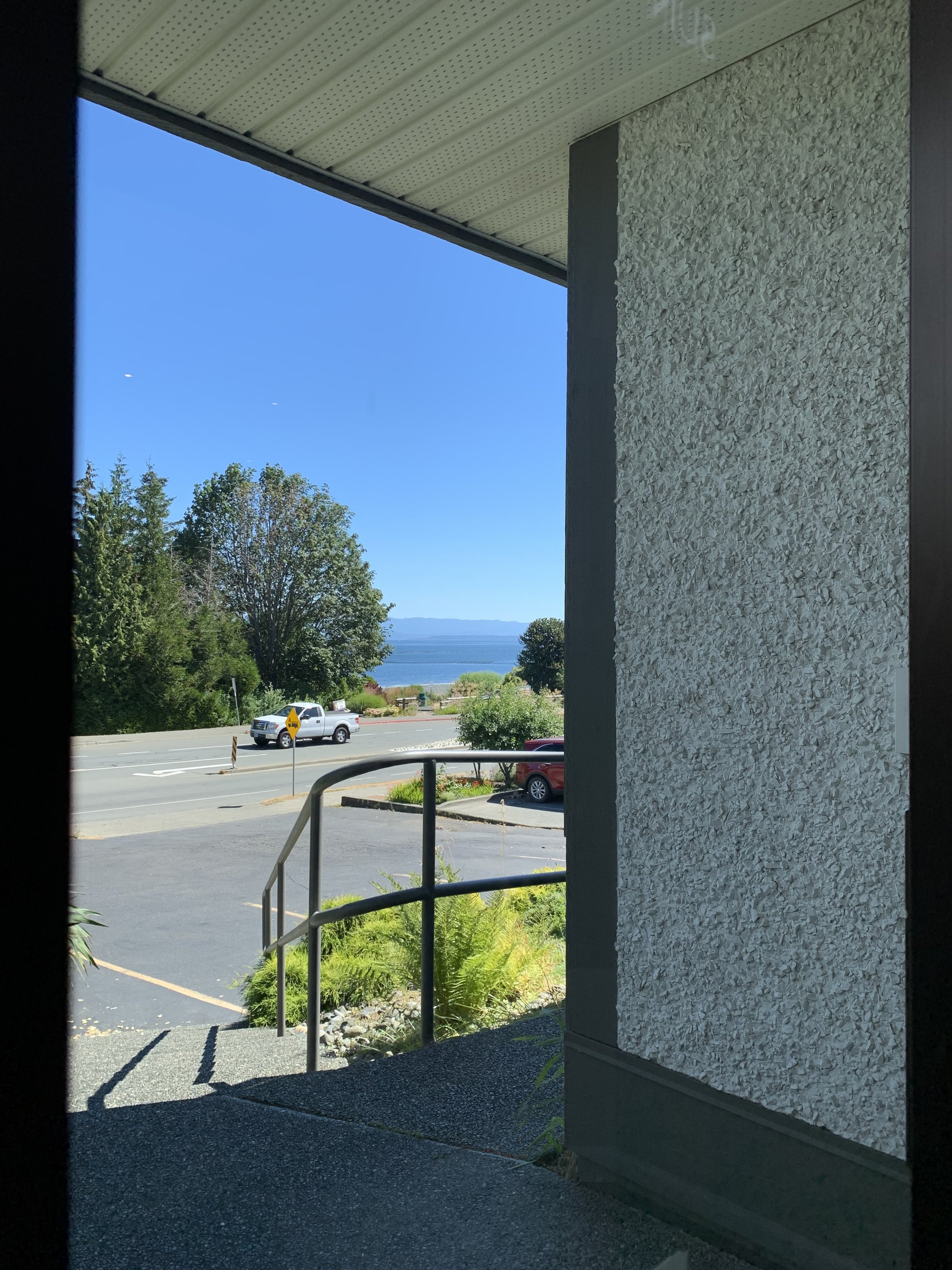 A view from inside a building looking out through an open doorway at a parking lot with a silver pickup truck, trees, a body of water, and a clear blue sky.