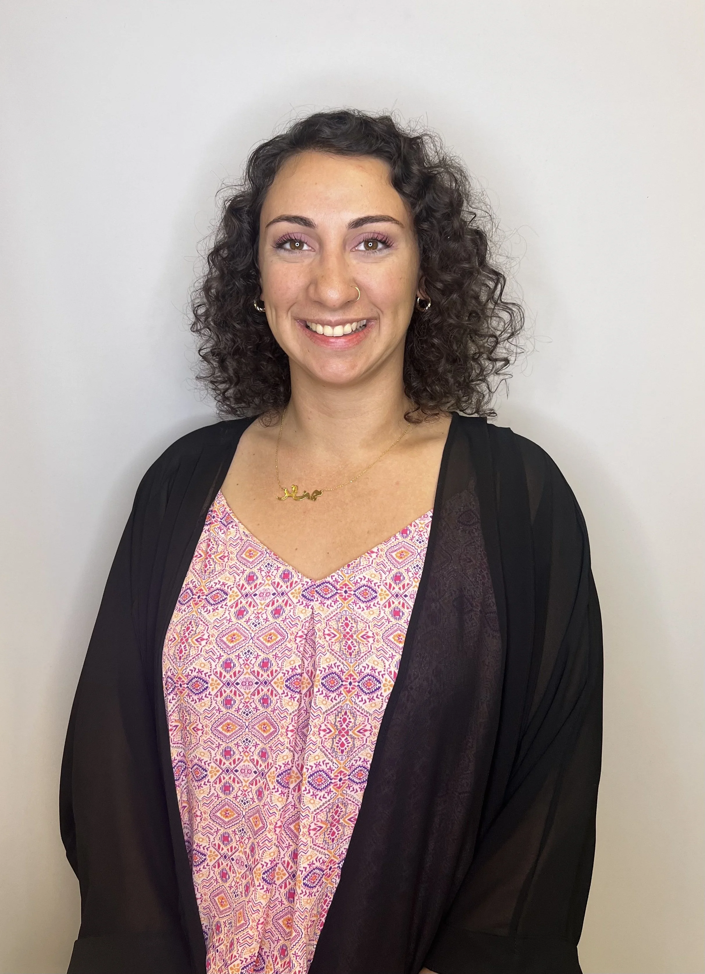 A smiling woman with curly dark hair wearing a pink patterned dress, a black cardigan, and gold jewelry, standing against a plain gray background.