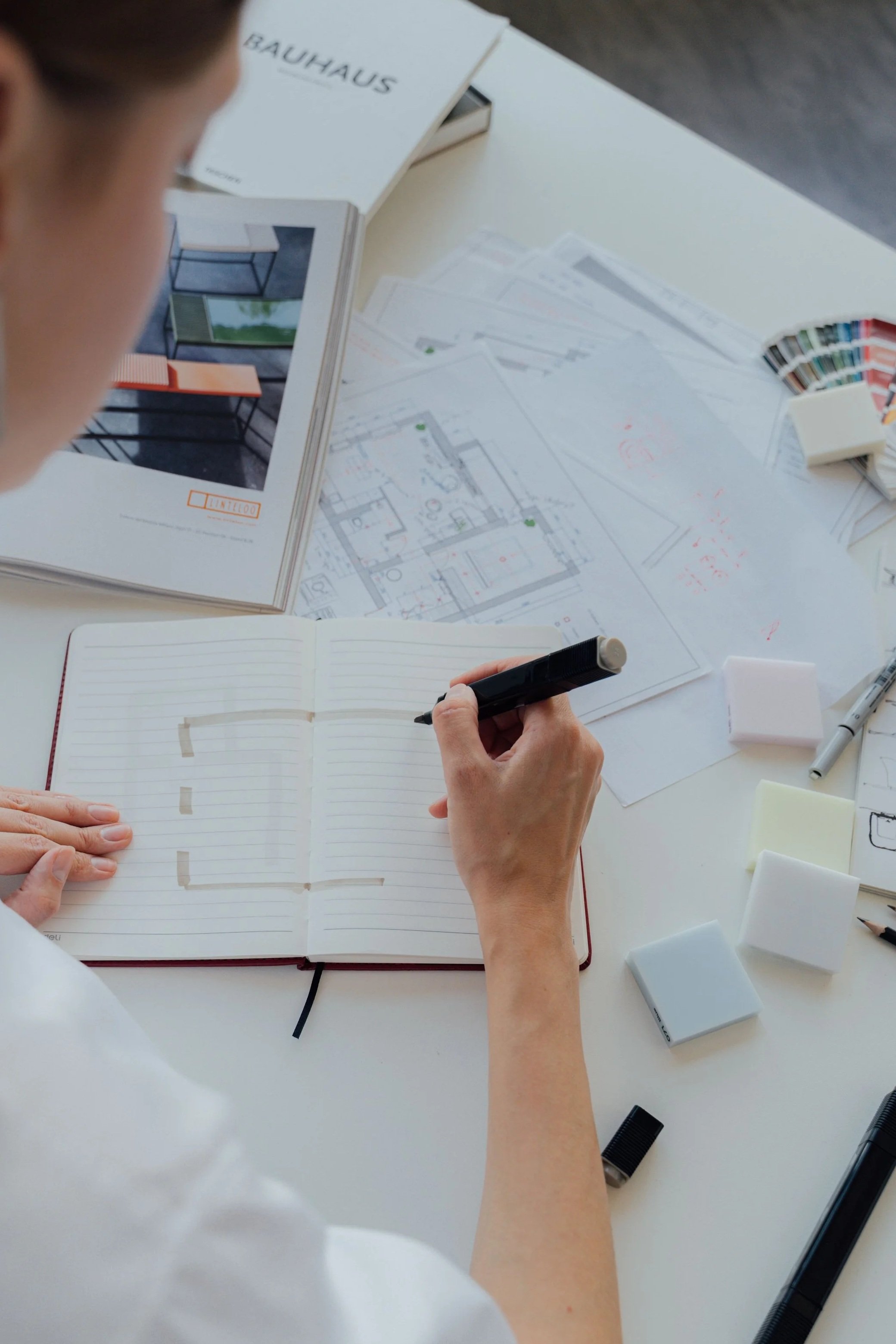 A person working on architectural plans with tools, color swatches, and a booklet on interior design. The person is holding a black marker and pointing to a notebook with sketches.