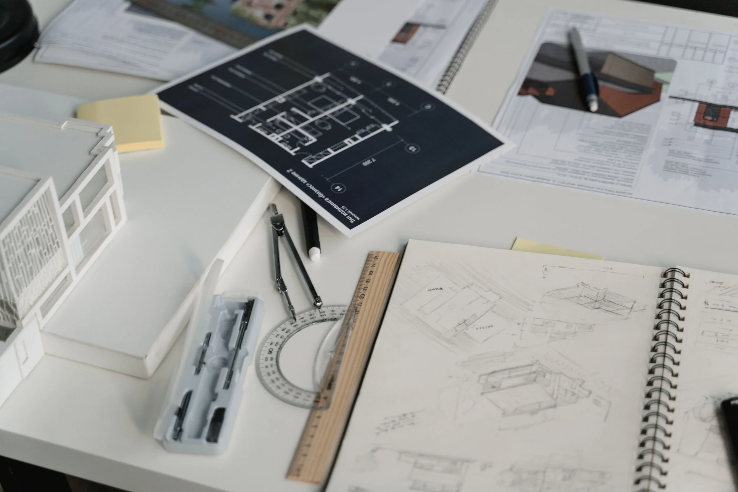 Architectural plans and blueprints spread out on a white table, with drafting tools like a ruler, protractor, and pens, along with a small model of a building.