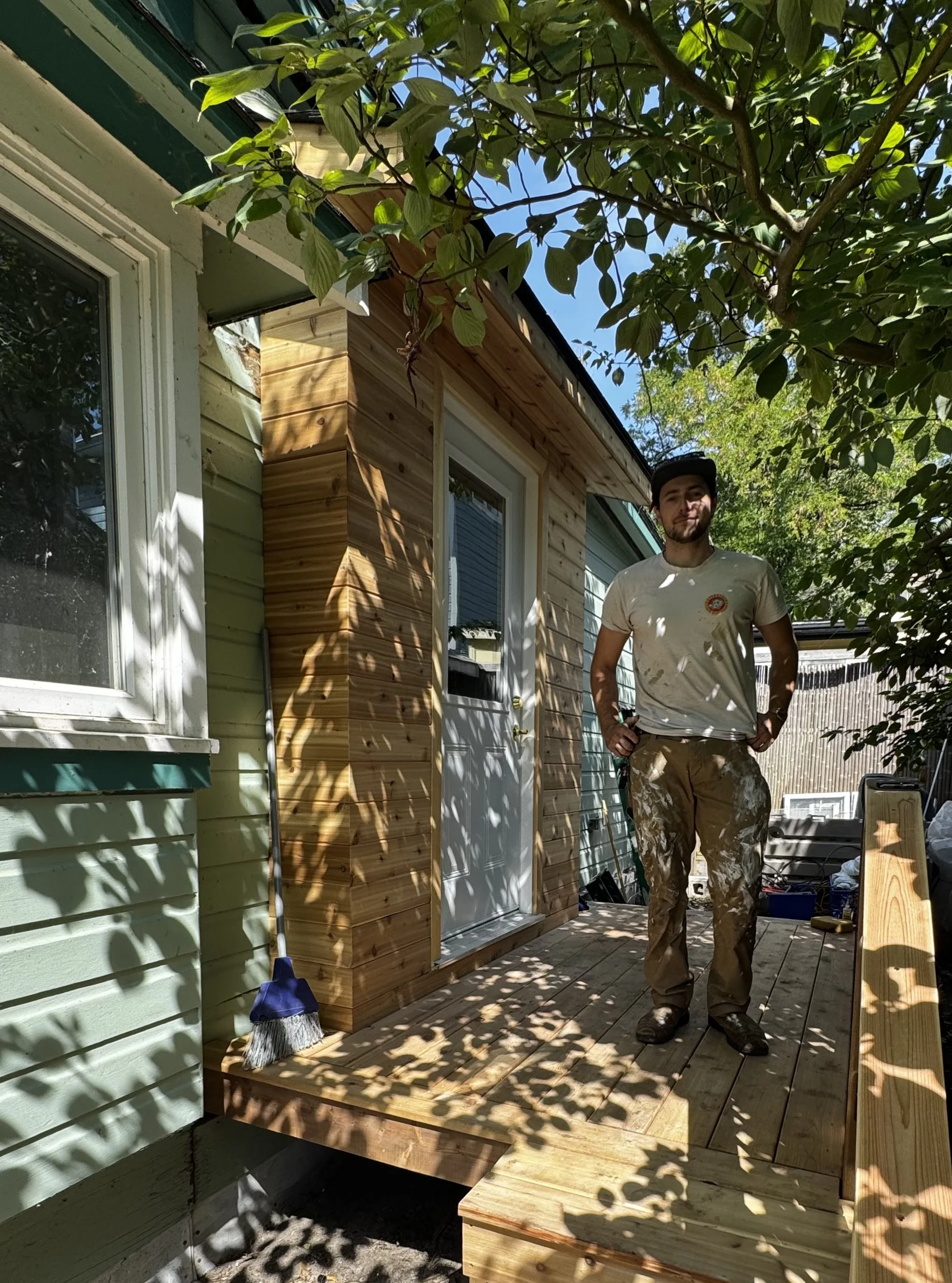 A man in work clothes standing outside on a wooden deck next to a house with new wooden siding and green painted house exterior, surrounded by trees and sunlight.