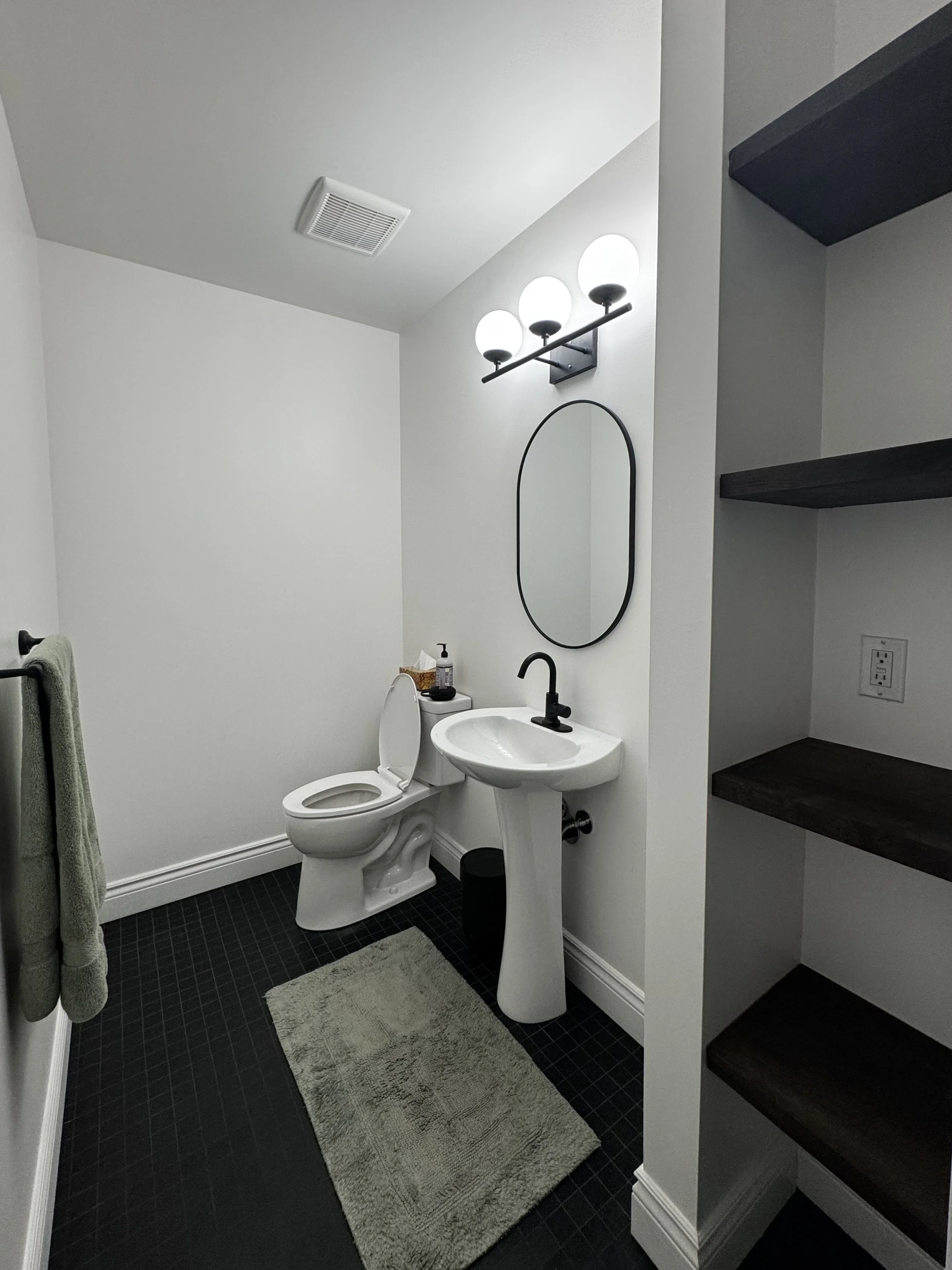Bathroom with a toilet, a small sink, a wall mirror, a light fixture with three globes, dark floor tiles, a beige rug, a towel on a towel rack, and dark wooden shelves on the wall.