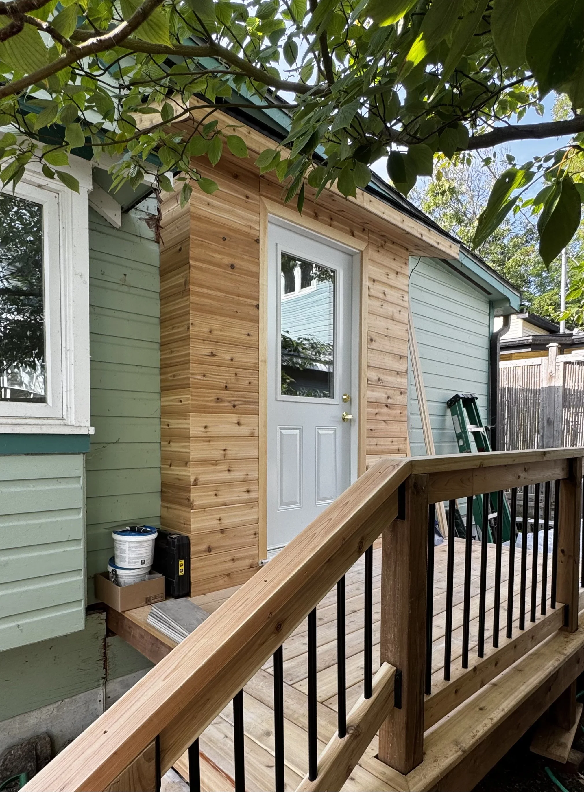 View of a house exterior undergoing home improvement, with wooden siding and a deck under construction, surrounded by trees and a wooden fence.