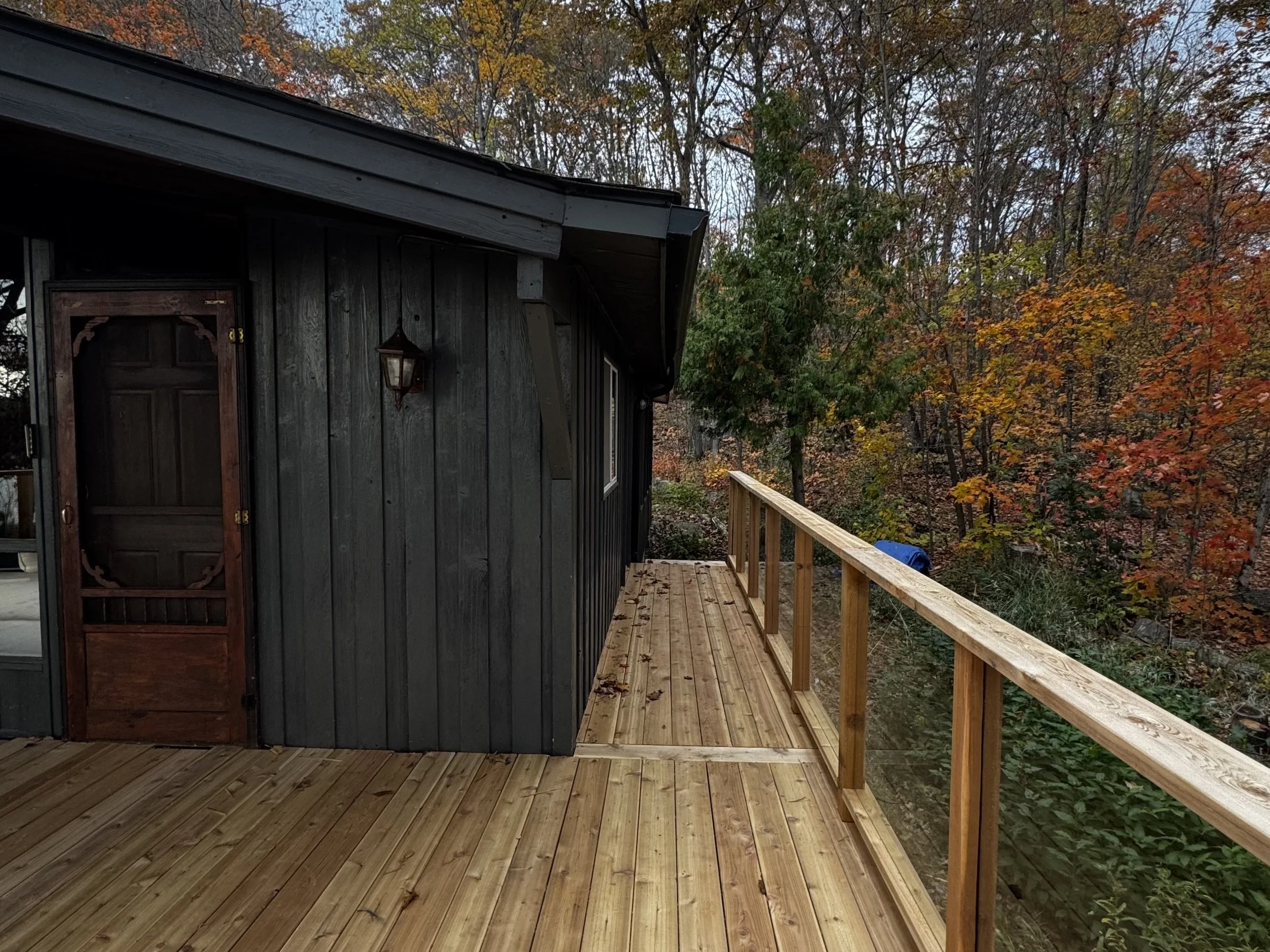 A wooden deck with a railing extends from a gray house with a dark brown door and a lantern outside. The background features trees with autumn foliage.