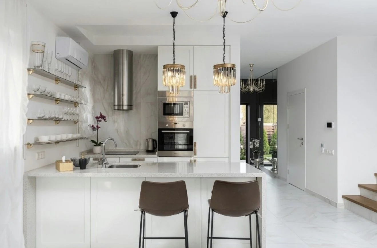 Modern kitchen with white cabinets and marble backsplash, open shelves with dishes, two brown chairs at kitchen island, stainless steel appliances, pendant and chandelier lighting, view of a patio through glass door.