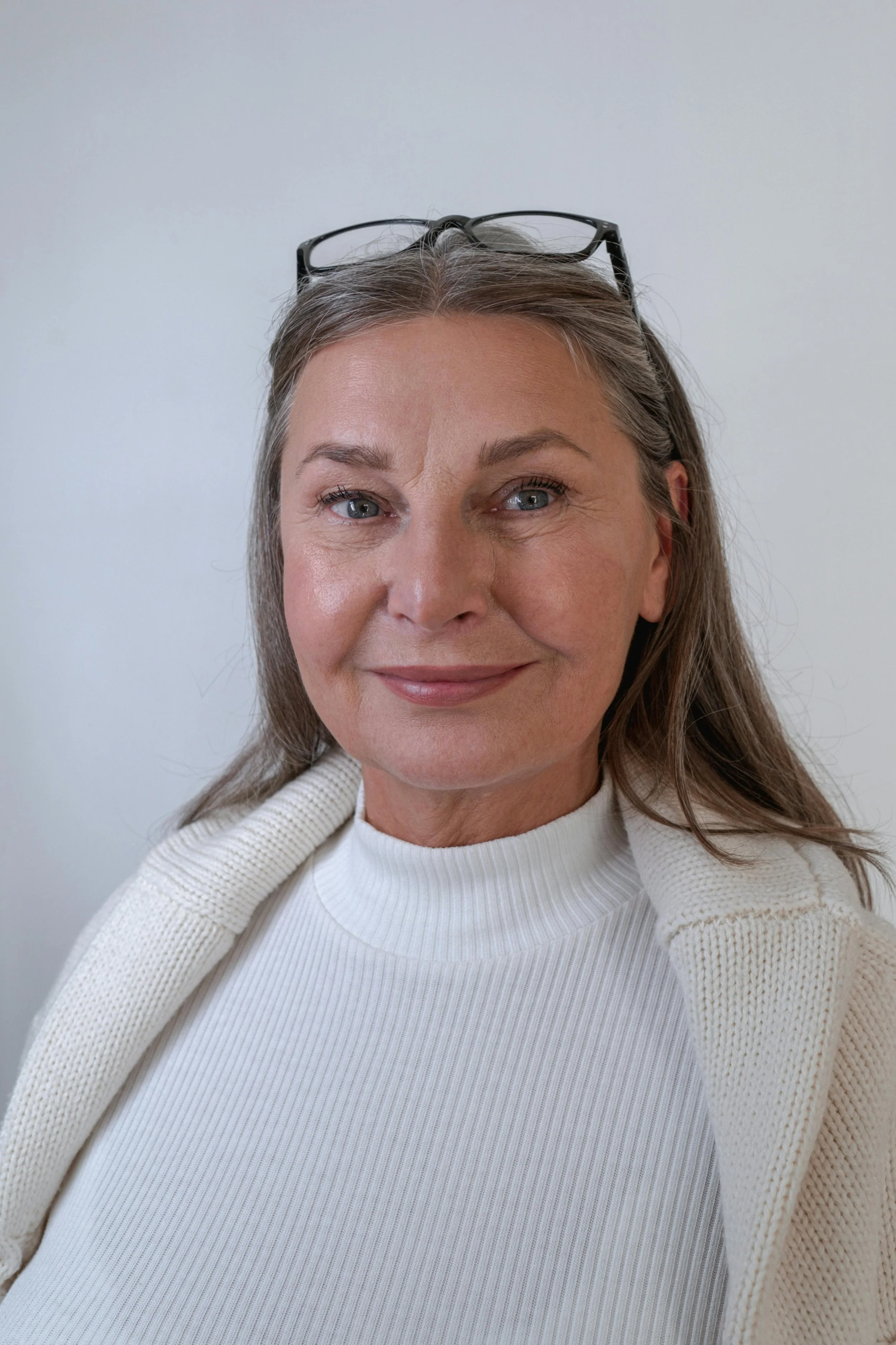 A smiling middle-aged woman with gray hair, blue eyes, and glasses resting on her head, wearing a white ribbed turtleneck sweater and a beige knit cardigan against a plain white wall.