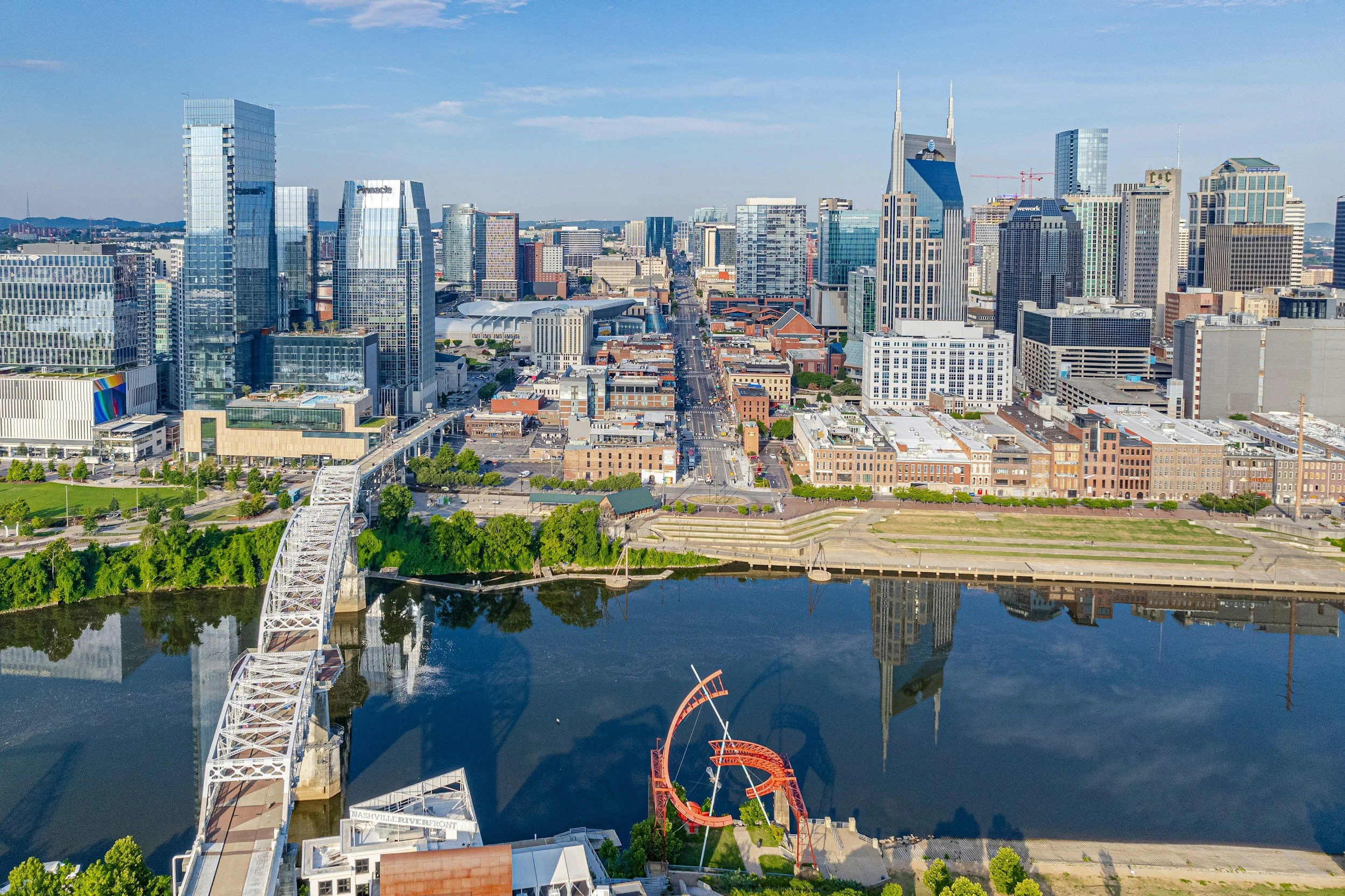 Aerial view of downtown Nashville with tall modern skyscrapers, the Cumberland River in the foreground, a white arched pedestrian bridge, and a red roller coaster nearby.