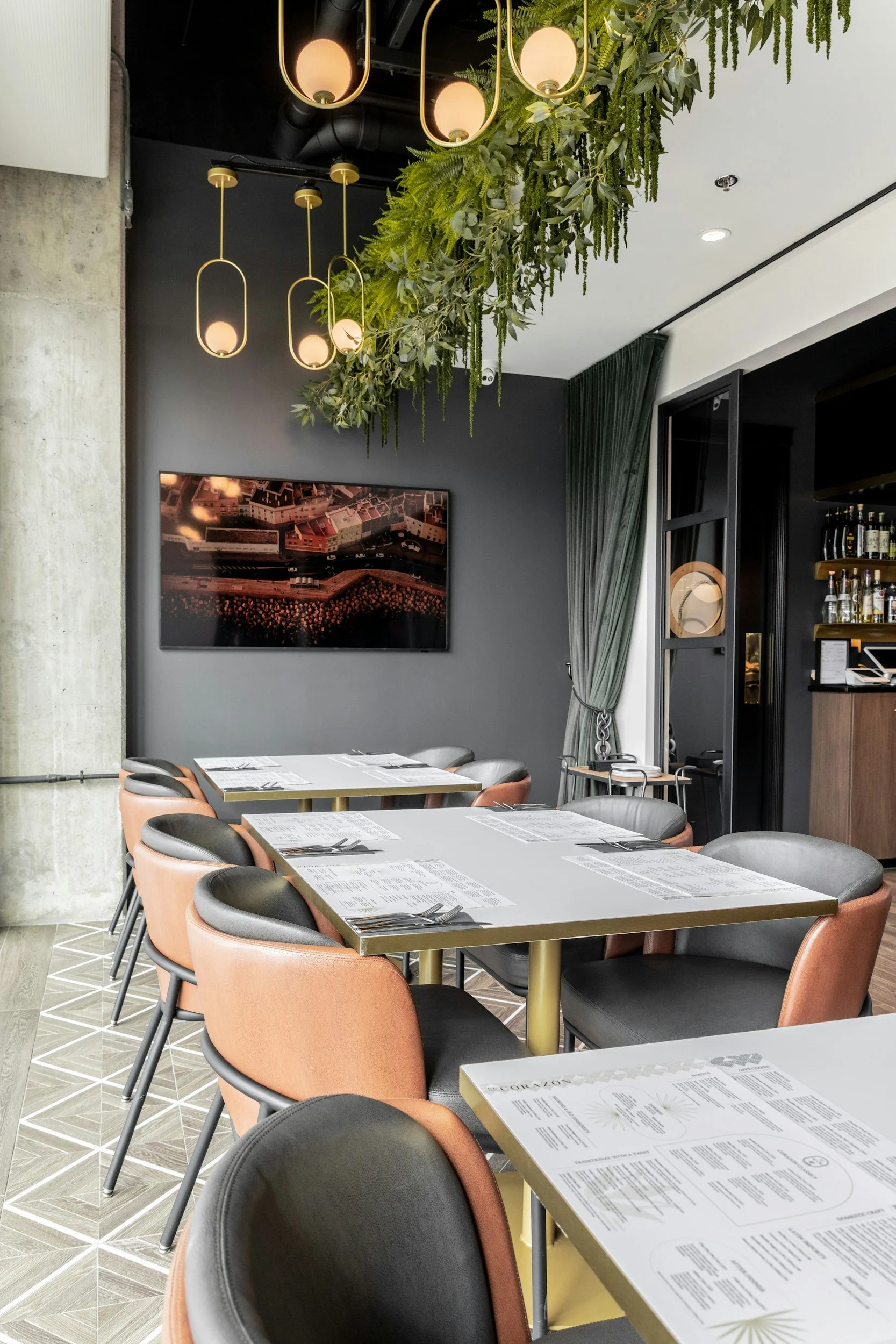 Interior of a modern restaurant with gray chairs featuring peach-colored upholstery, tables set with menus and utensils, a dark gray wall with a landscape photograph, lush green plants hanging from the ceiling, and contemporary light fixtures with round bulbs.