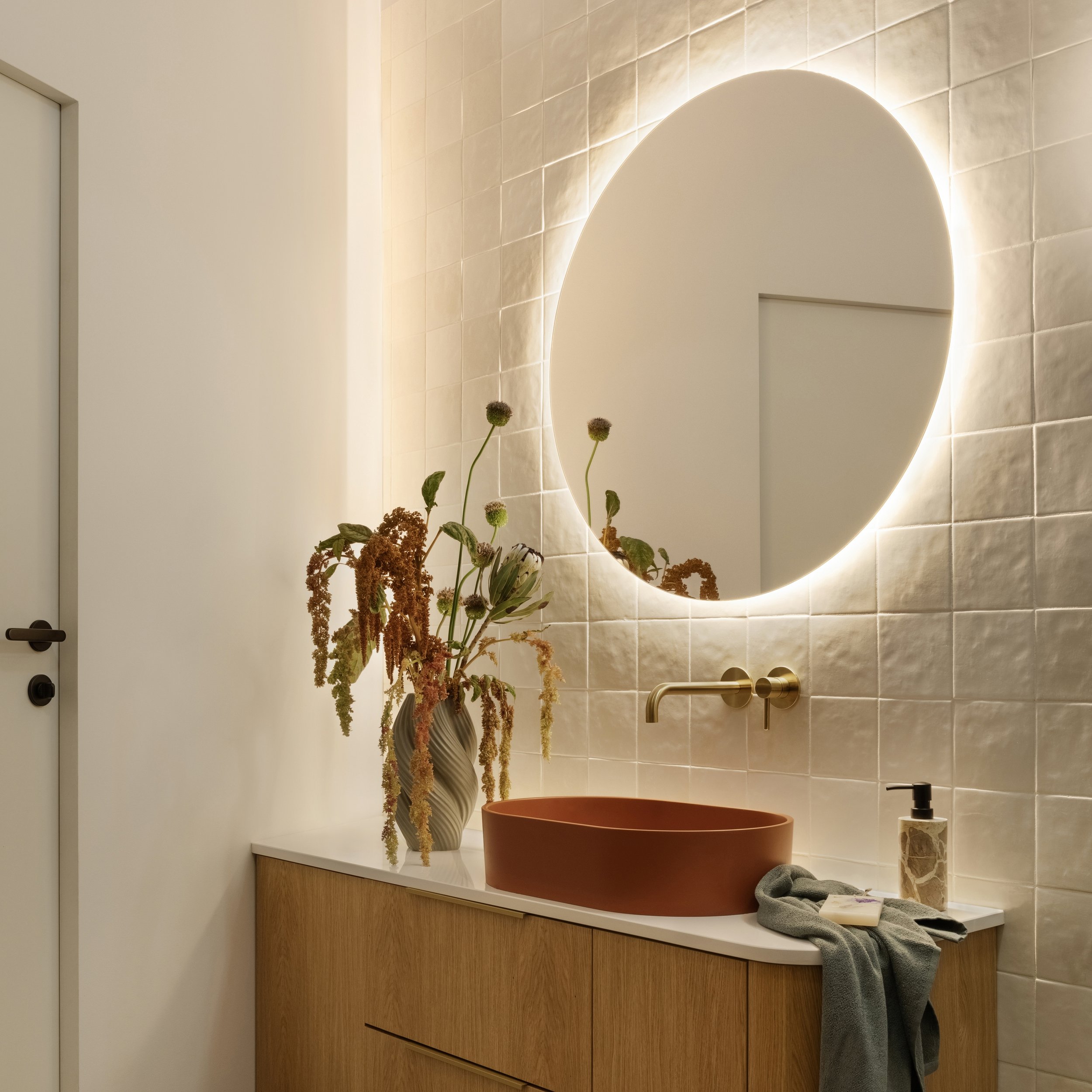 A modern bathroom vanity with a large mirror illuminated by backlighting, a vase with dried flowers, a terracotta-colored basin, and a gray towel on the countertop. The wall behind has beige tiles.