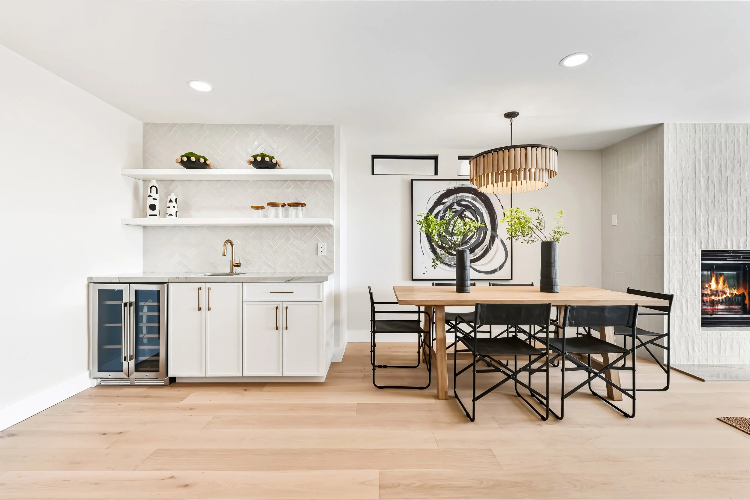Modern dining room with a light wood dining table, black chairs, a decorative chandelier, a large abstract black and white wall art, a fireplace, and a built-in kitchen with white cabinets, open shelving, and black and white decor.