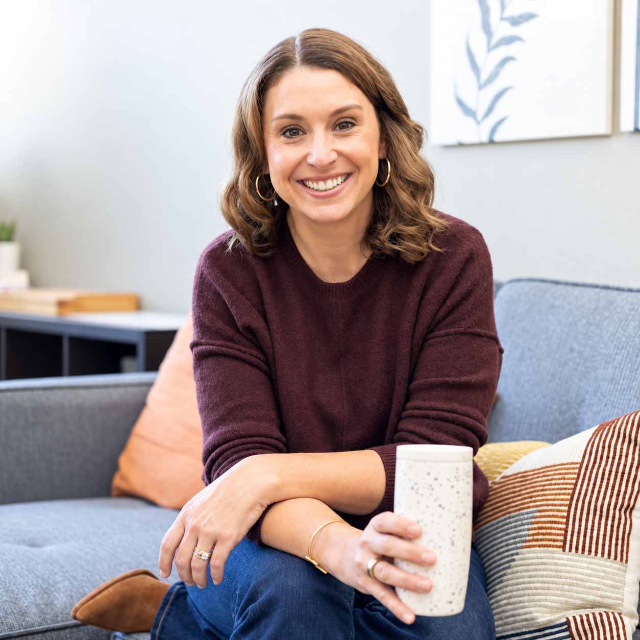 A woman with shoulder-length brown hair and hoop earrings smiling while sitting on a sofa, holding a speckled white mug.