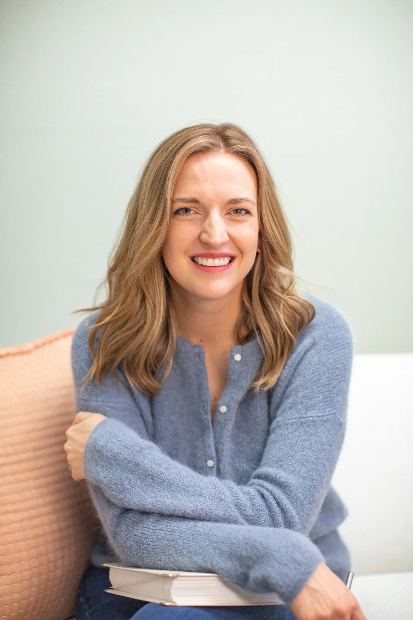 A woman with wavy, light brown hair and fair skin smiling, sitting with her arms crossed, wearing a blue cardigan, in a room with soft green walls and a light-colored chair.