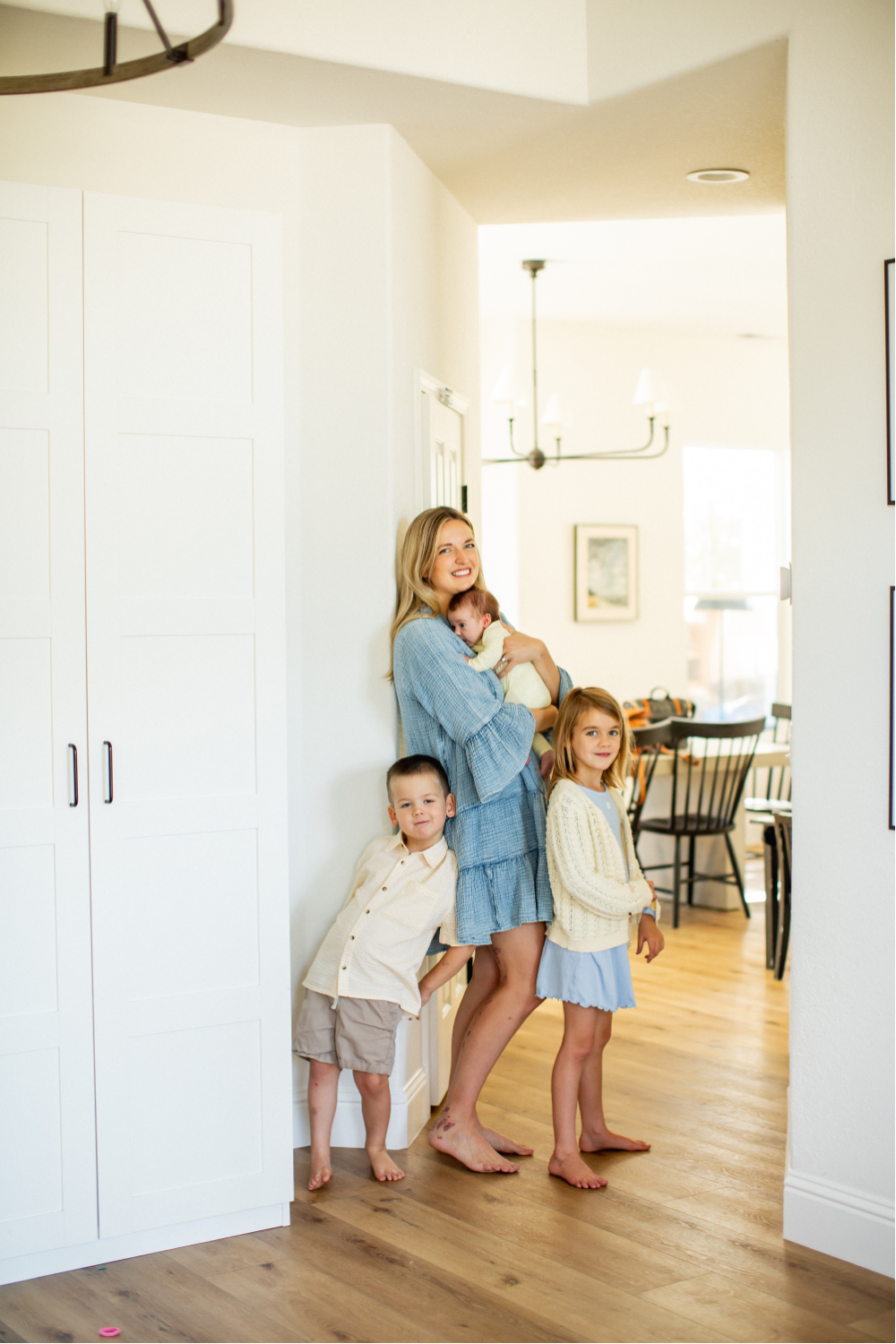 A woman with blonde hair holding a baby, standing with two children in a bright home interior.
