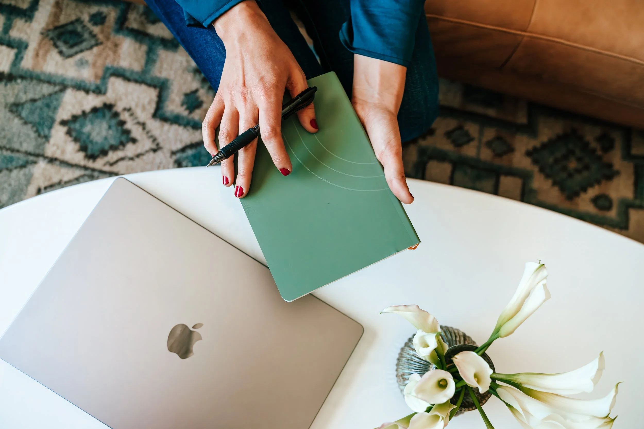 A person with red nail polish writing on a green folder with a black pen, sitting beside an Apple iMac computer and a vase of white calla lilies.