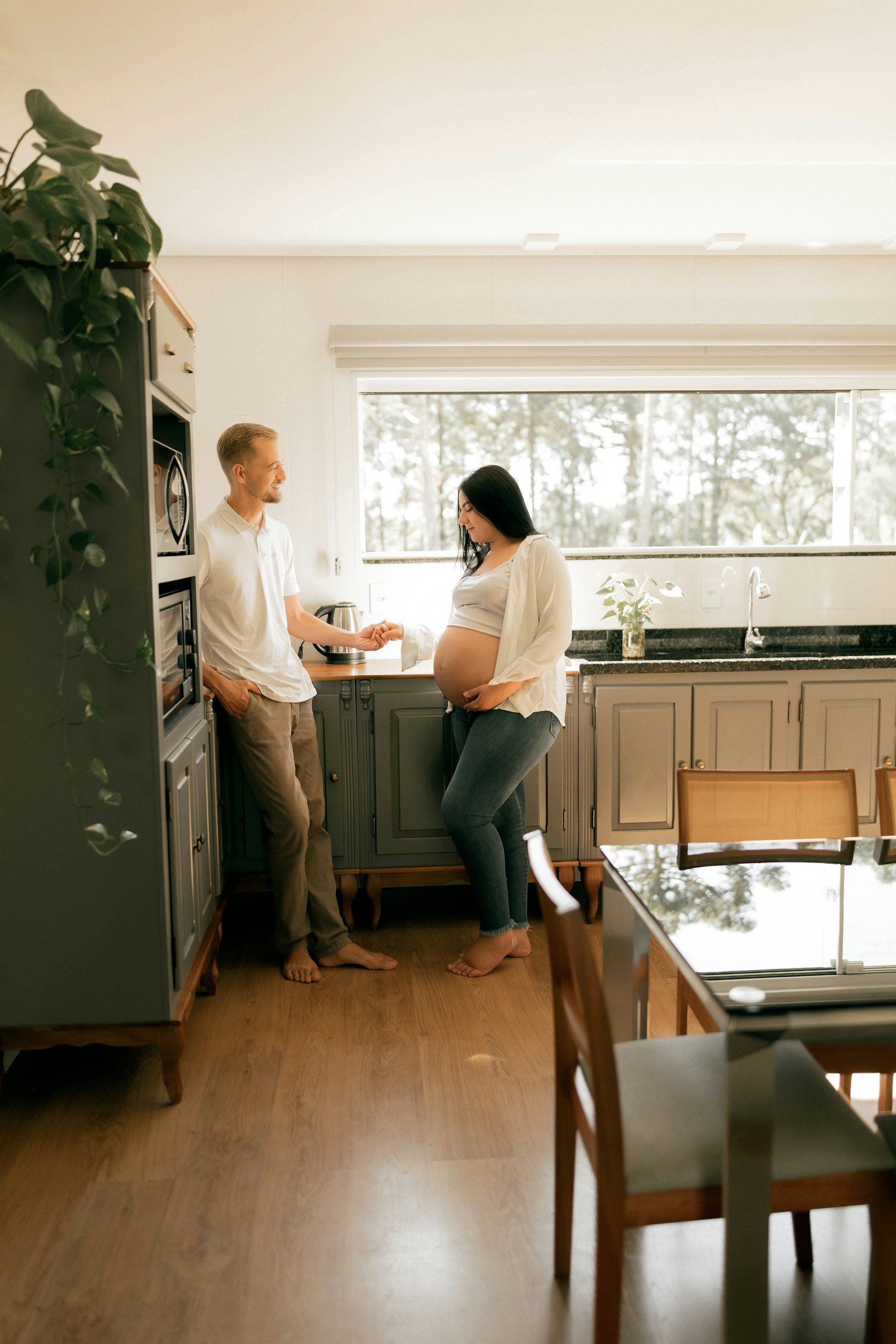 Pregnant woman and man with hand on her belly, standing in a kitchen, smiling and holding hands, with a window showing a forest outside.