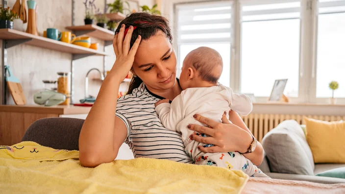 A woman sitting at a table holding a small baby in her arms, looking tired or stressed in a cozy home kitchen and dining area.
