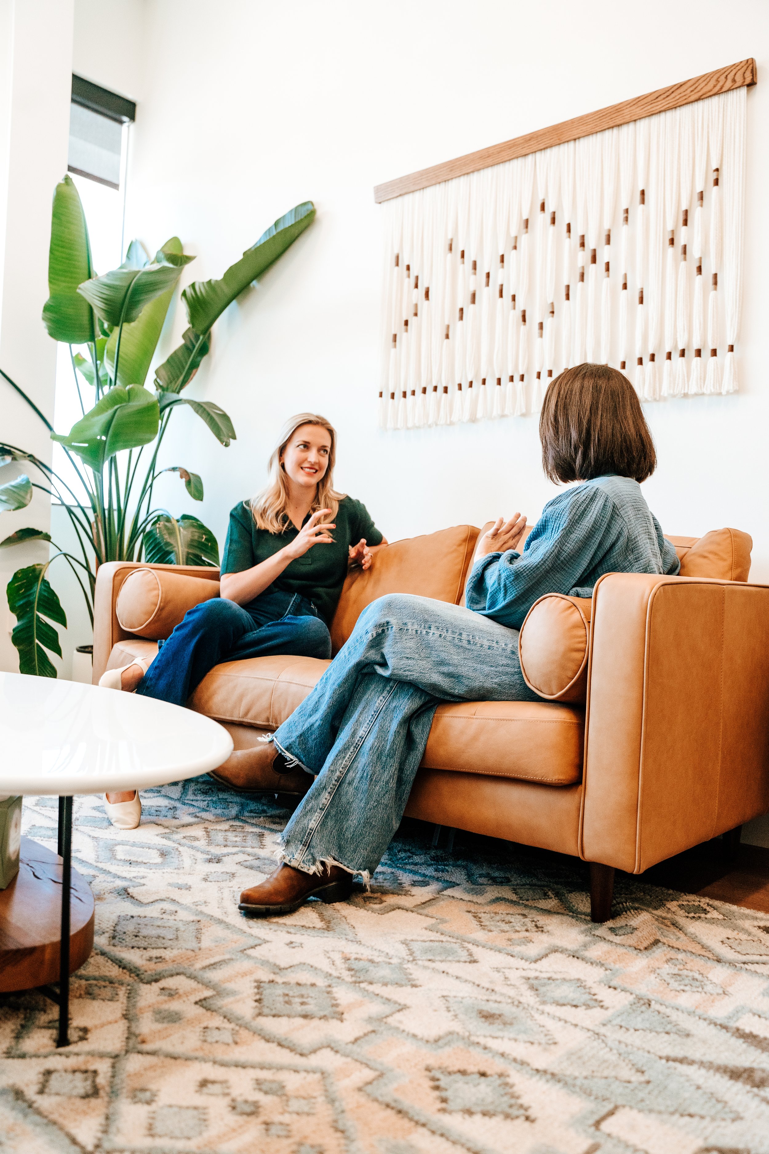 Two women are sitting on a tan sofa in a living room, engaging in conversation. One woman has blonde hair, is wearing a black top, and gesturing with her hands. The other has brown hair, is wearing a gray top, and is listening. There is a large green plant to the left and a decorative woven wall hanging above them. A white coffee table is partially visible, and the floor is covered with a patterned rug.