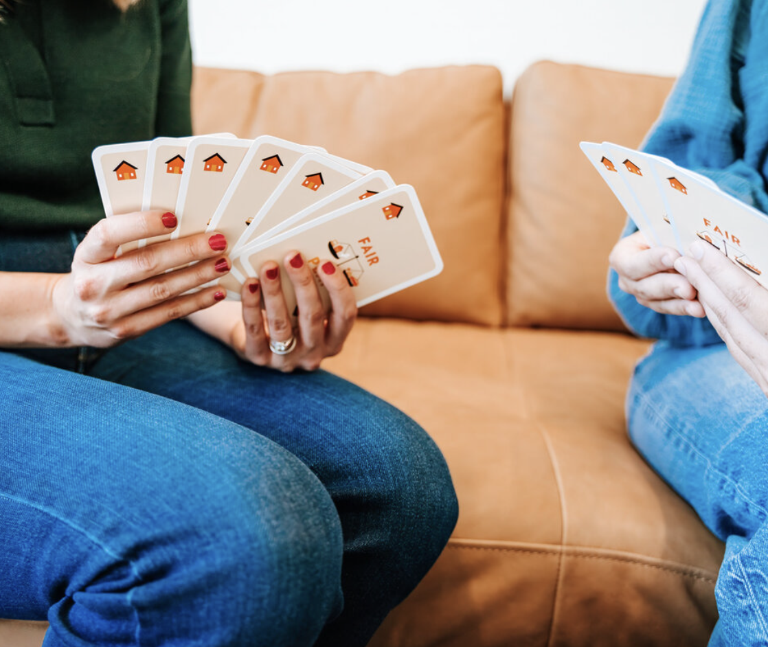 Two people playing a card game on a couch, holding cards with a house logo and the words 'Fair Deal'.
