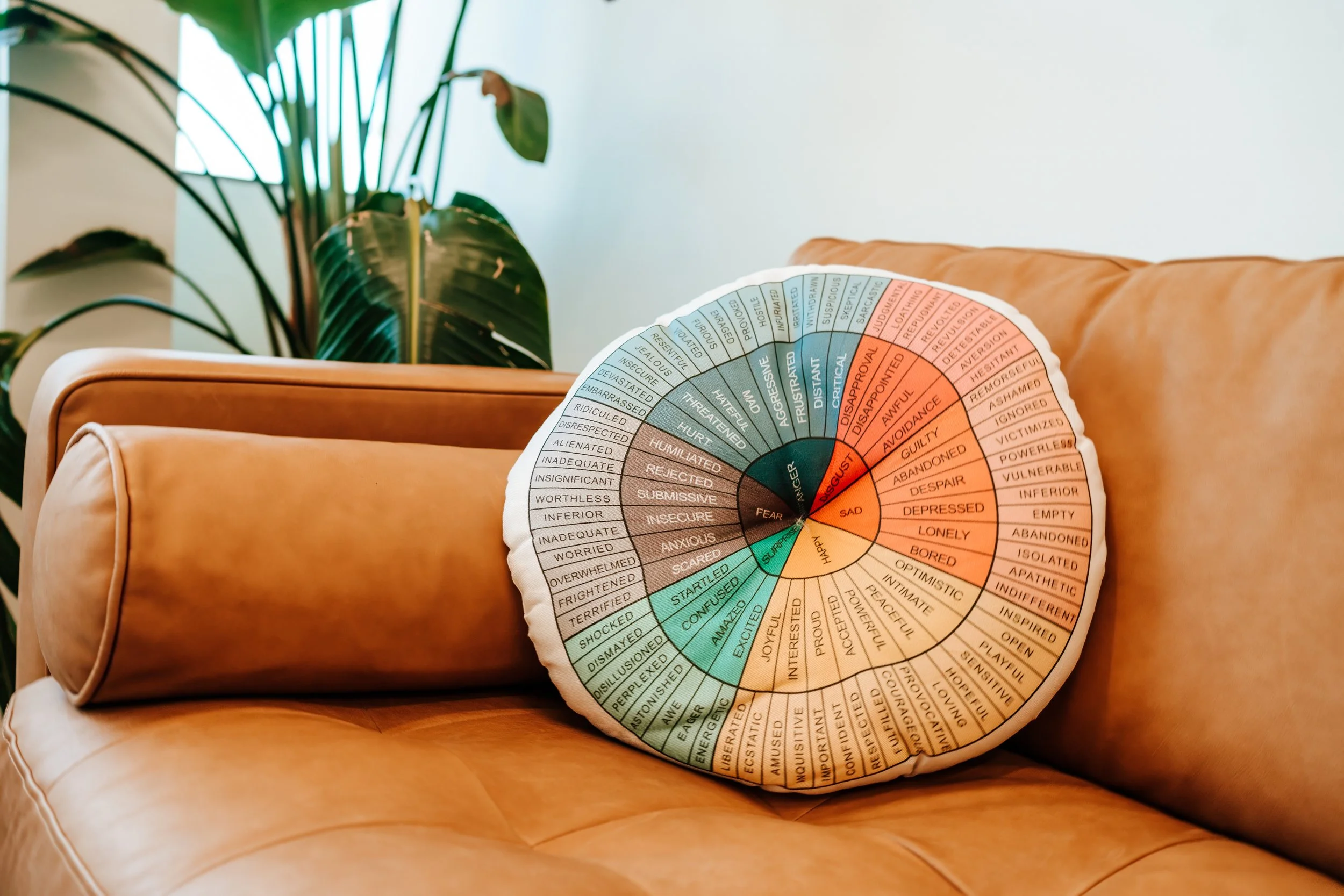 A round emotion wheel pillow resting on a tan leather couch, with a large green leafy plant in the background.