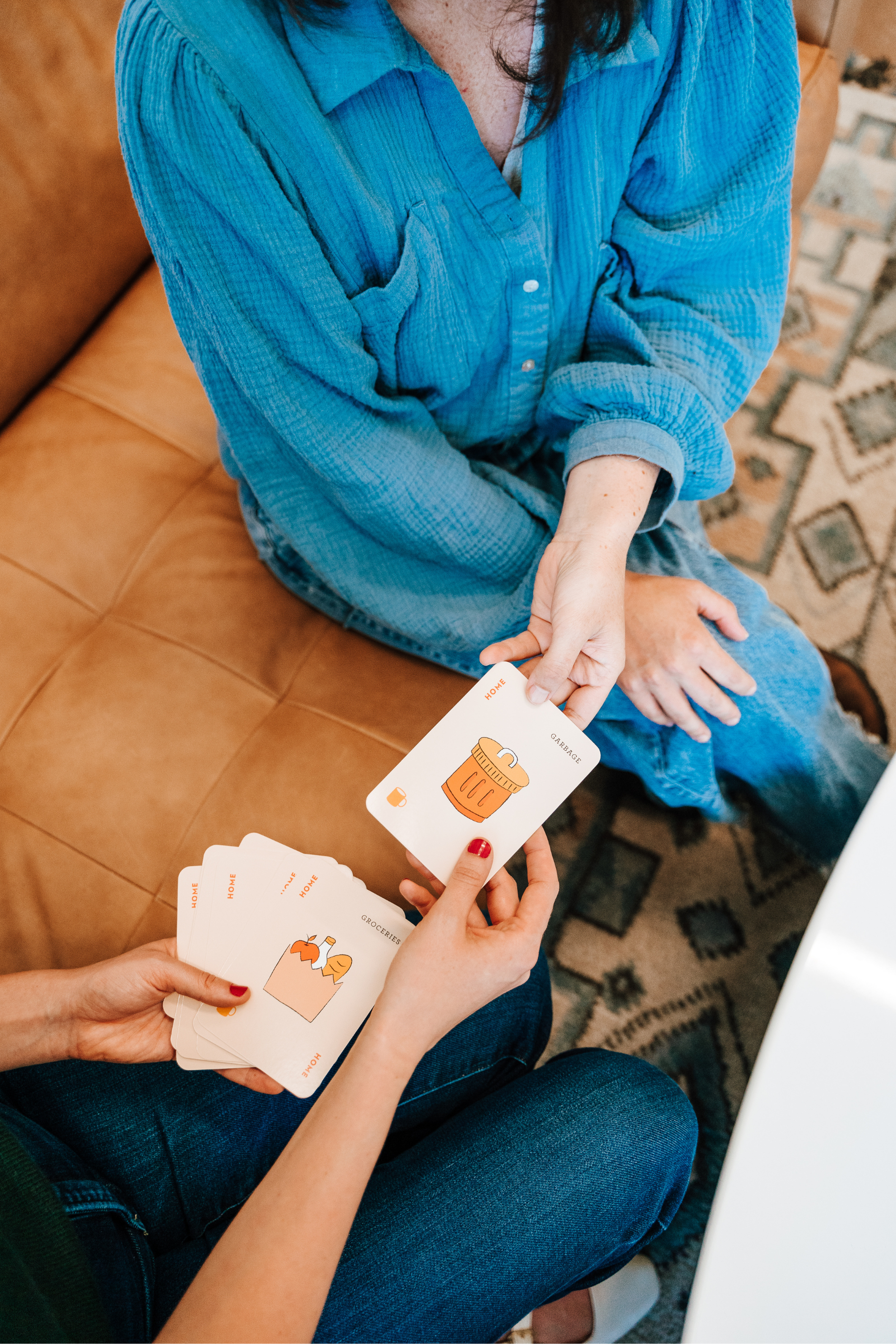 People playing a card game with illustrated cards, sitting on a brown leather couch with a patterned rug nearby.