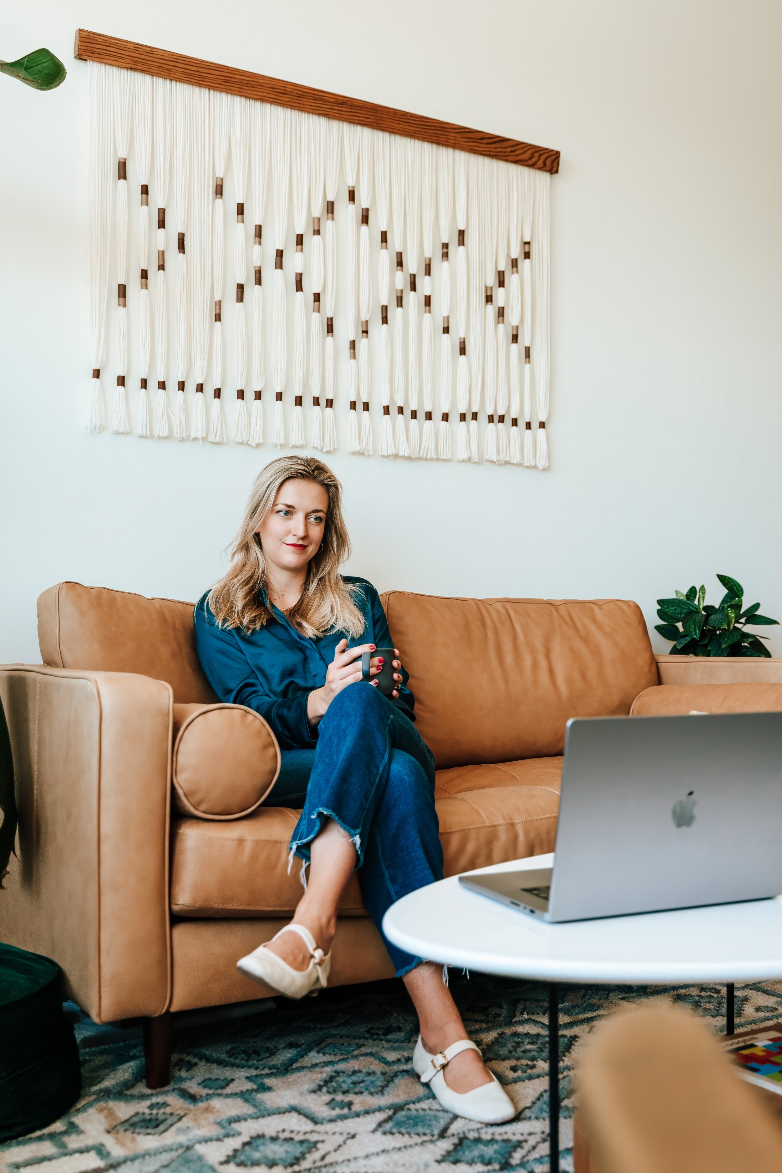 A woman with blonde hair sitting on a tan sofa, holding a mug, with a laptop on a white coffee table in front of her, in a living room with wall art, a green plant, and a patterned rug.