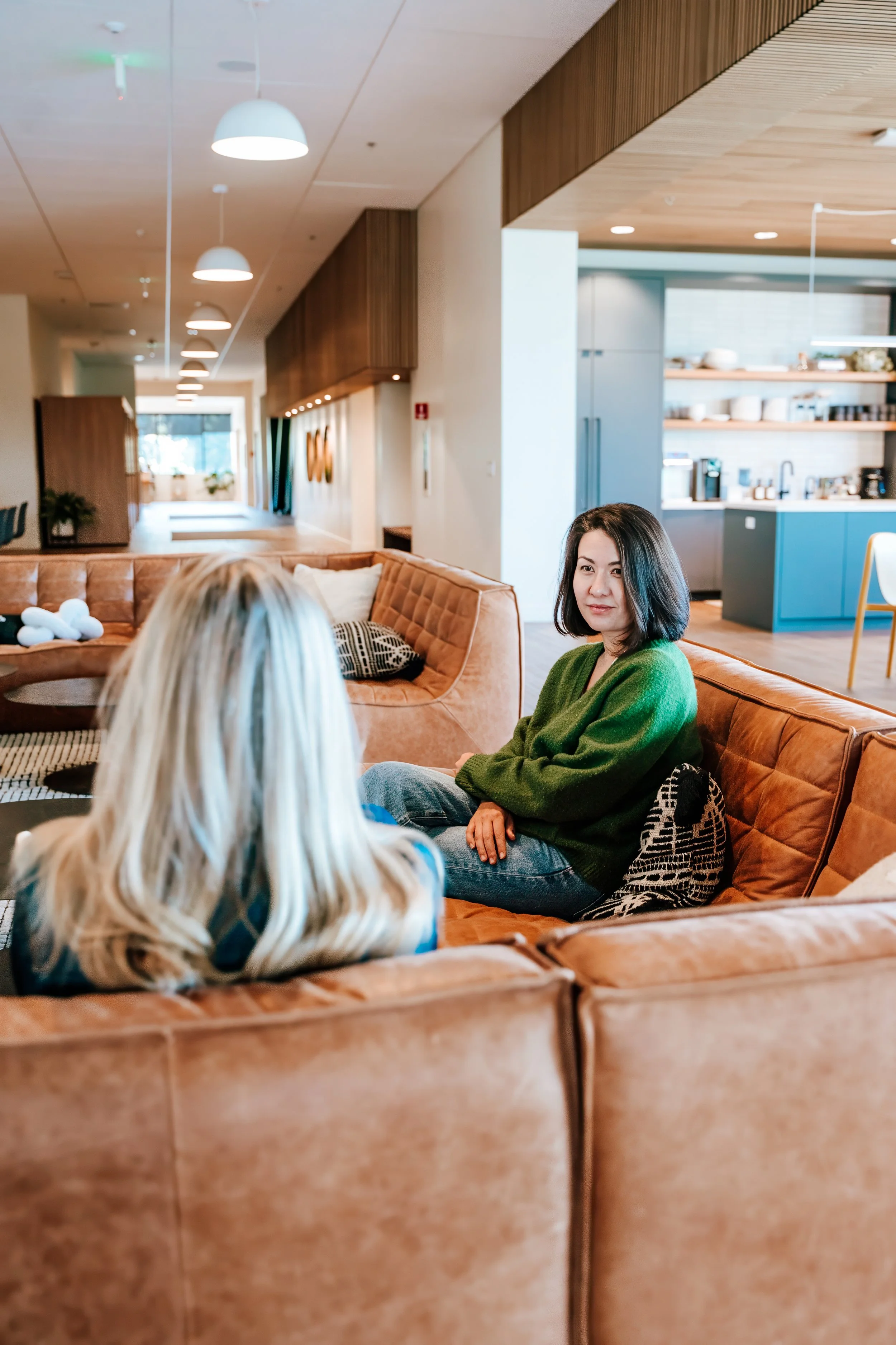 Two women sitting on a light brown couch in a modern living room; one woman with dark hair wearing a green sweater, the other with blonde hair with her back facing the camera.