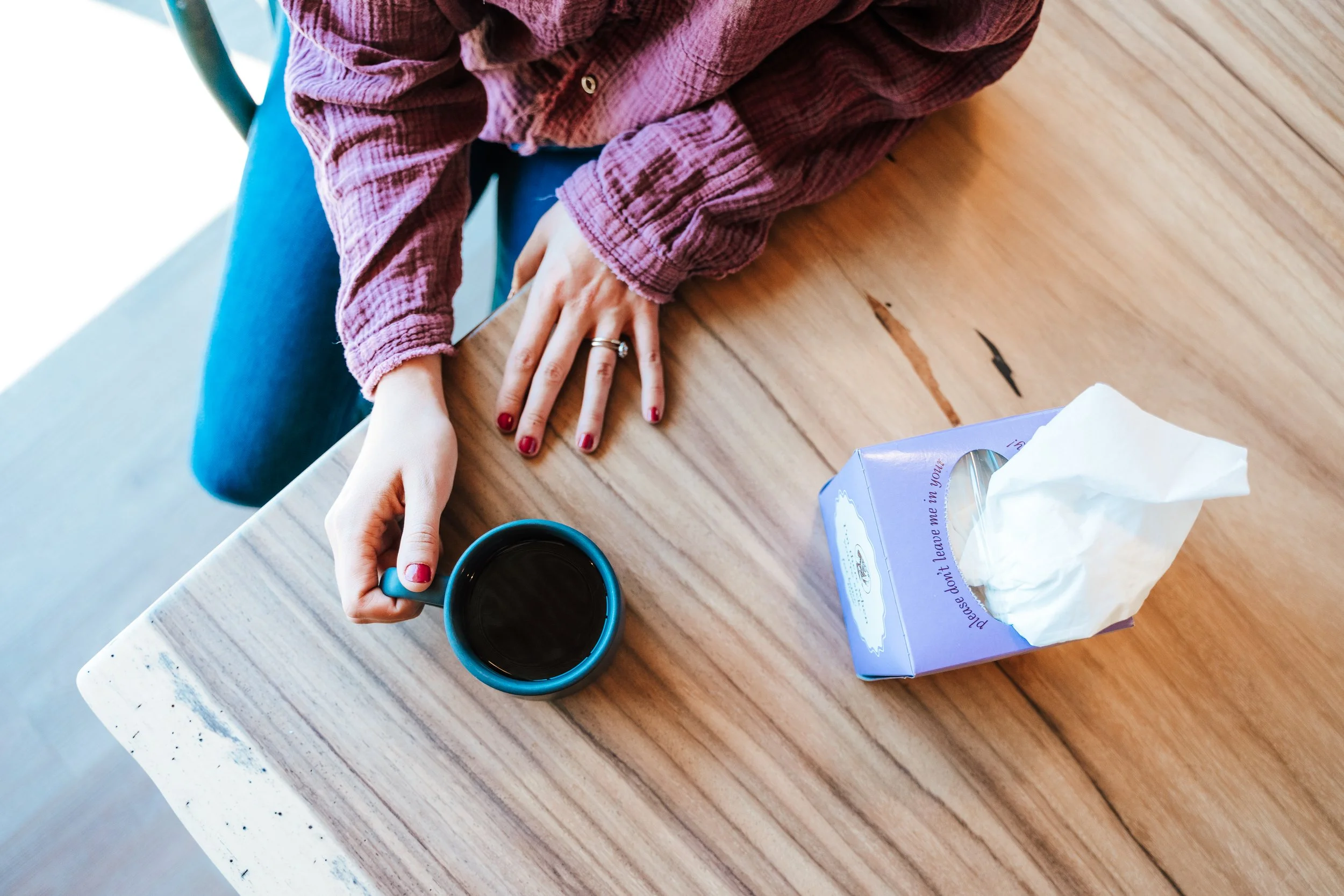 A person with painted nails and rings on their fingers is sitting at a wooden table, holding a cup of coffee, with a tissue box and a mug on the table.