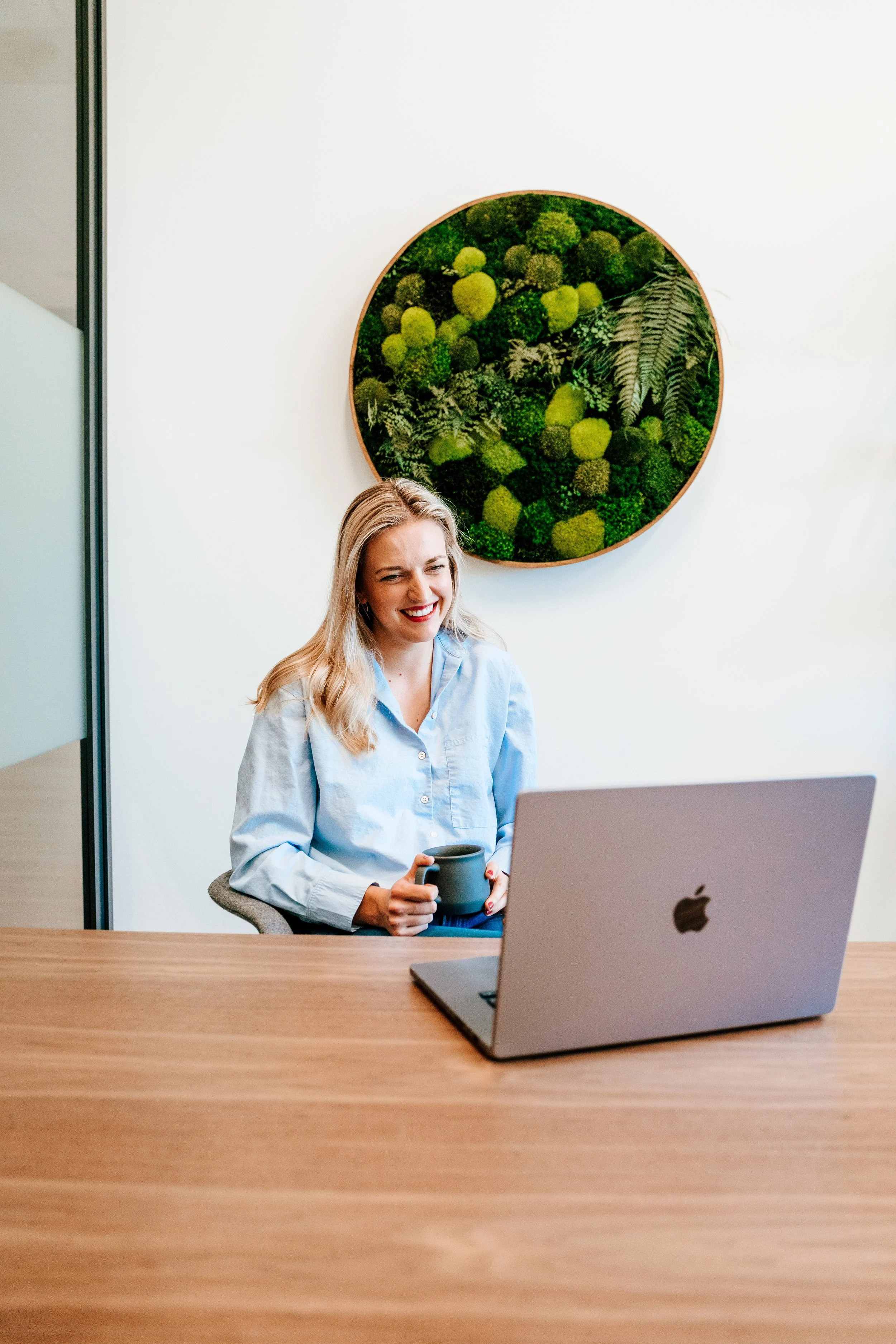 A woman sitting at a desk in an office, smiling, holding a mug, with a laptop and a desktop monitor on the desk. A circular wall art featuring green plants is visible on the wall behind her.