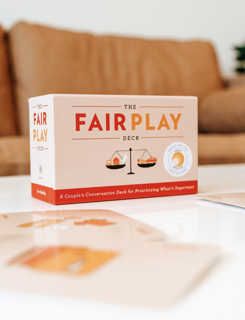The Fair Play deck box, designed for couples to prioritize important conversations, is placed on a white table with a blurred brown sofa and potted plant in the background.